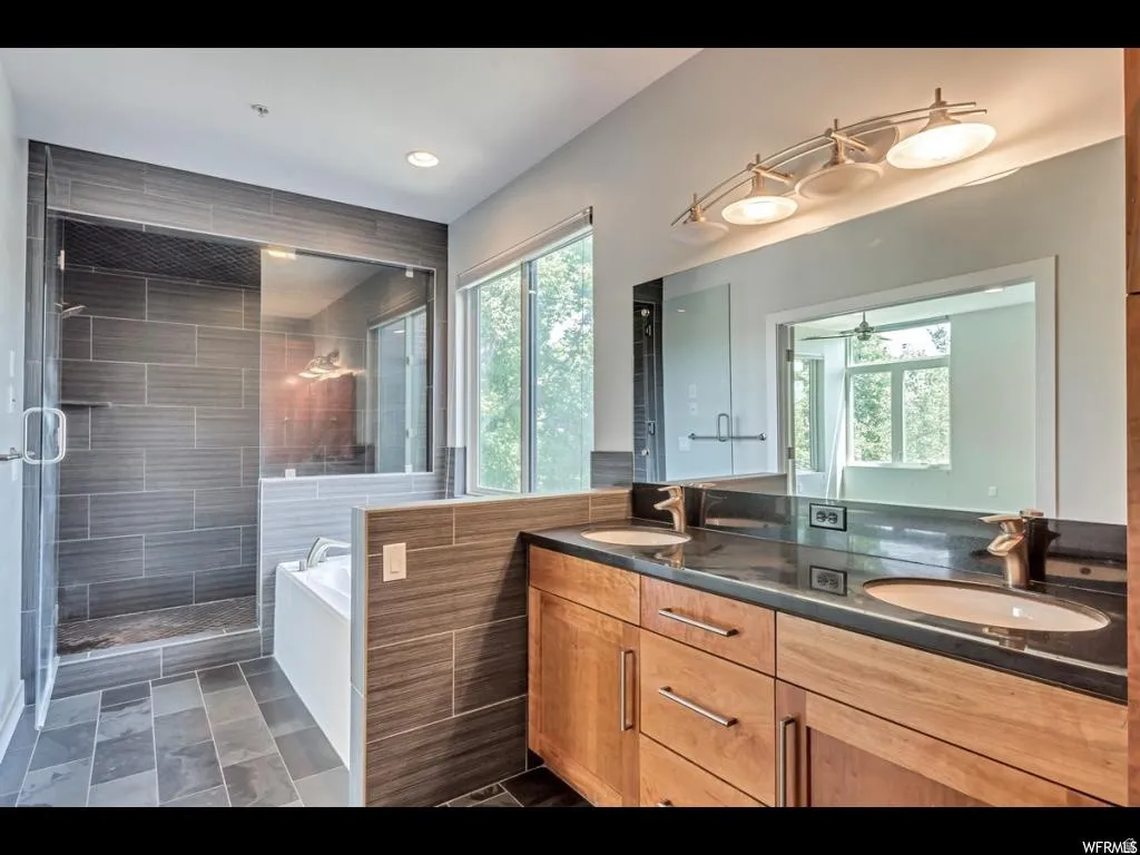 Spacious bathroom featuring a dual vanity with light wood cabinetry and dark stone countertop, a large mirror with overhead lighting, a deep soaking tub, and a separate glass-enclosed shower with dark gray tile surround and flooring