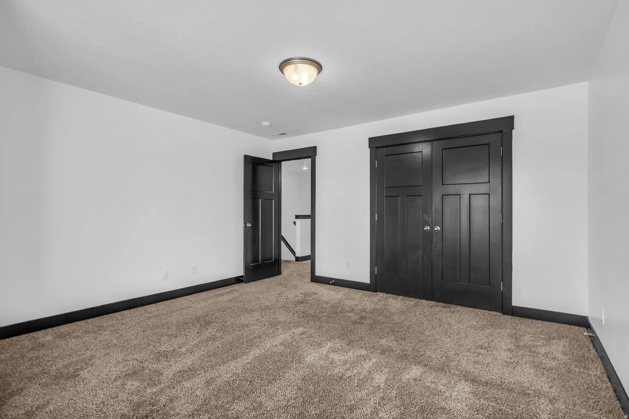 Spacious room featuring neutral carpeting, white walls, dark trim, dark paneled doors, and a flush-mount ceiling light fixture