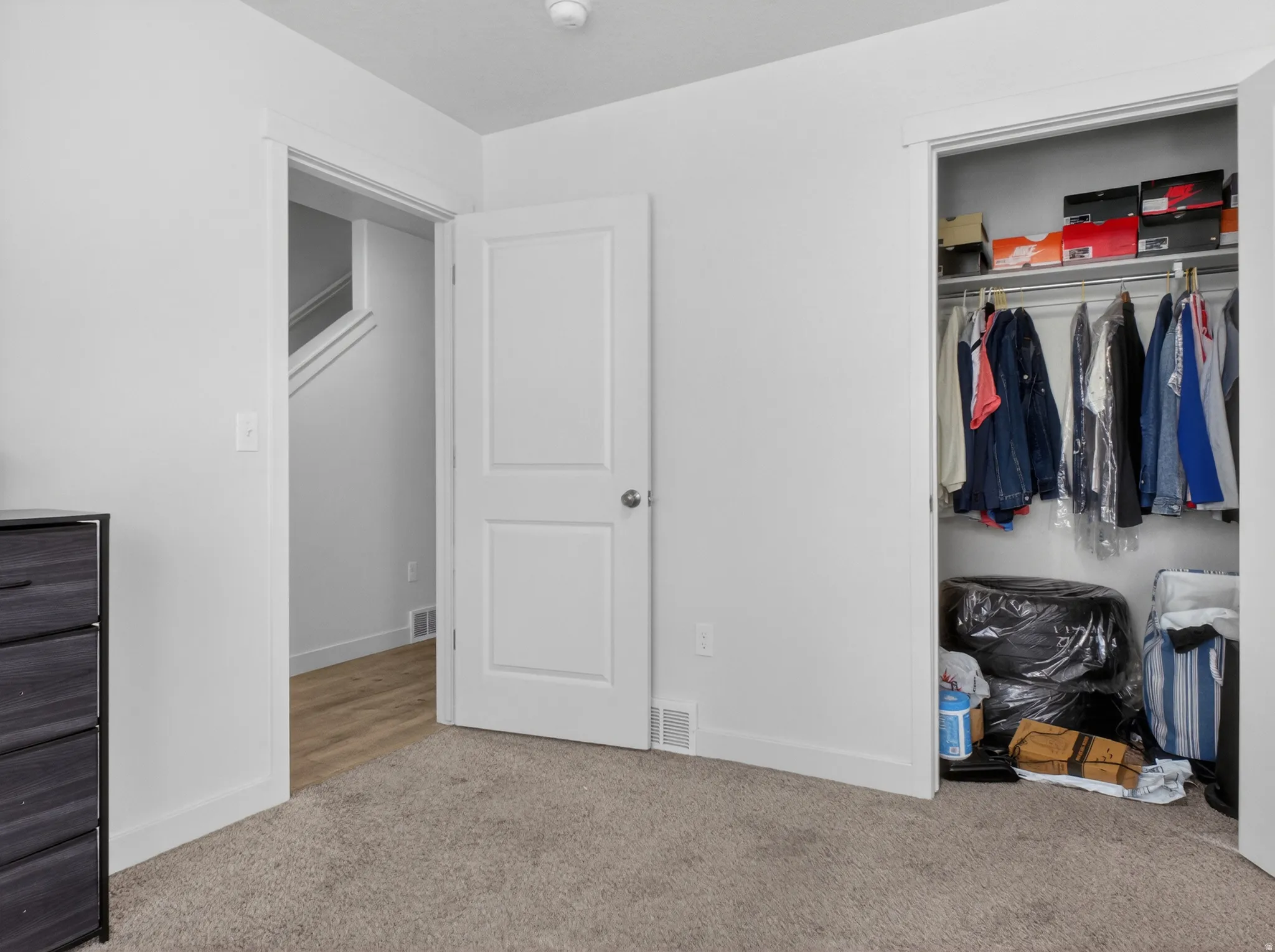 Carpeted interior space featuring a built-in closet with shelving and hanging rod, a white interior door, and light wood-finish flooring visible through an open doorway