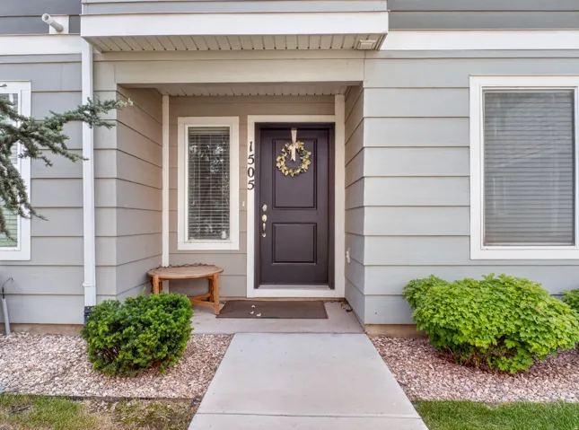Front entry featuring a dark-paneled door with house numbers, light gray horizontal siding, white trim windows, concrete pathway, and decorative gravel landscaping with green shrubbery