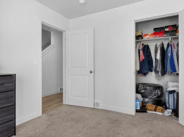 Carpeted interior space featuring a built-in closet with shelving and hanging rod, a white interior door, and light wood-finish flooring visible through an open doorway