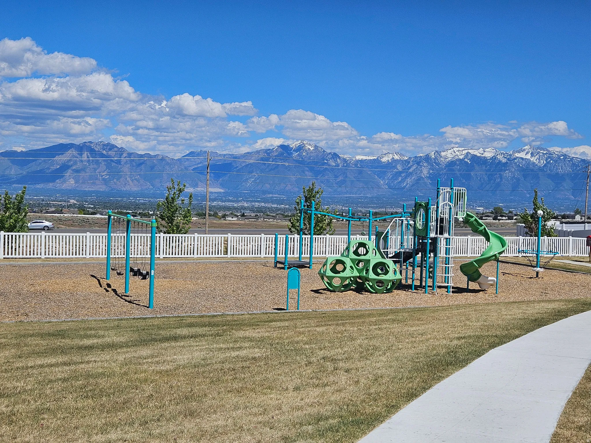 Community playground featuring green and blue play structures, a slide, and swing sets on a wood chip surface