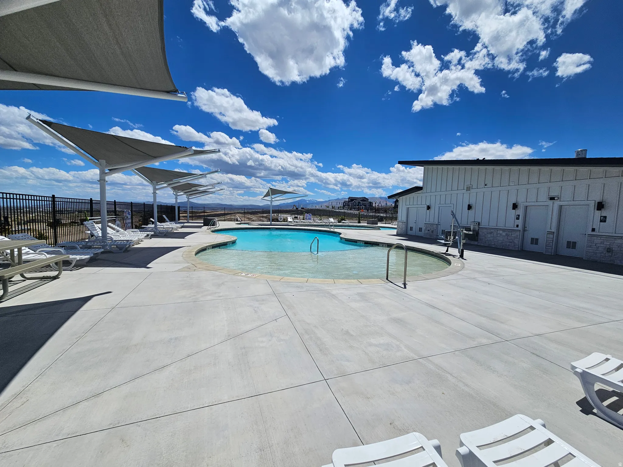 Outdoor community pool area featuring a curved swimming pool with a shallow entry, extensive concrete decking, multiple shade structures, and a dedicated pool house with vertical siding and stone accents