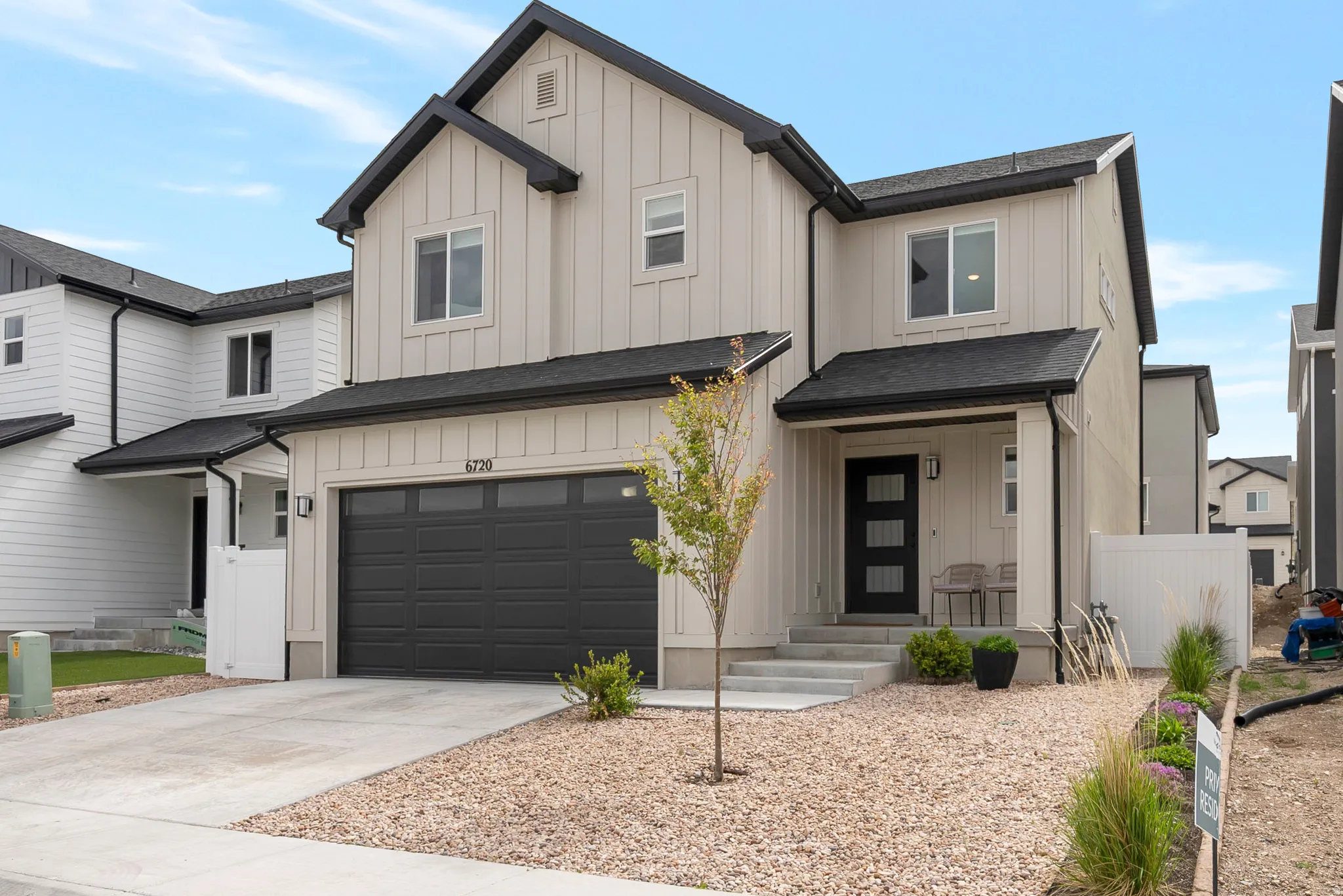 Modern two-story residence featuring light-toned vertical siding with dark trim accents, a two-car garage, covered entry porch, dark front door with frosted glass panels, and a low-maintenance gravel front yard