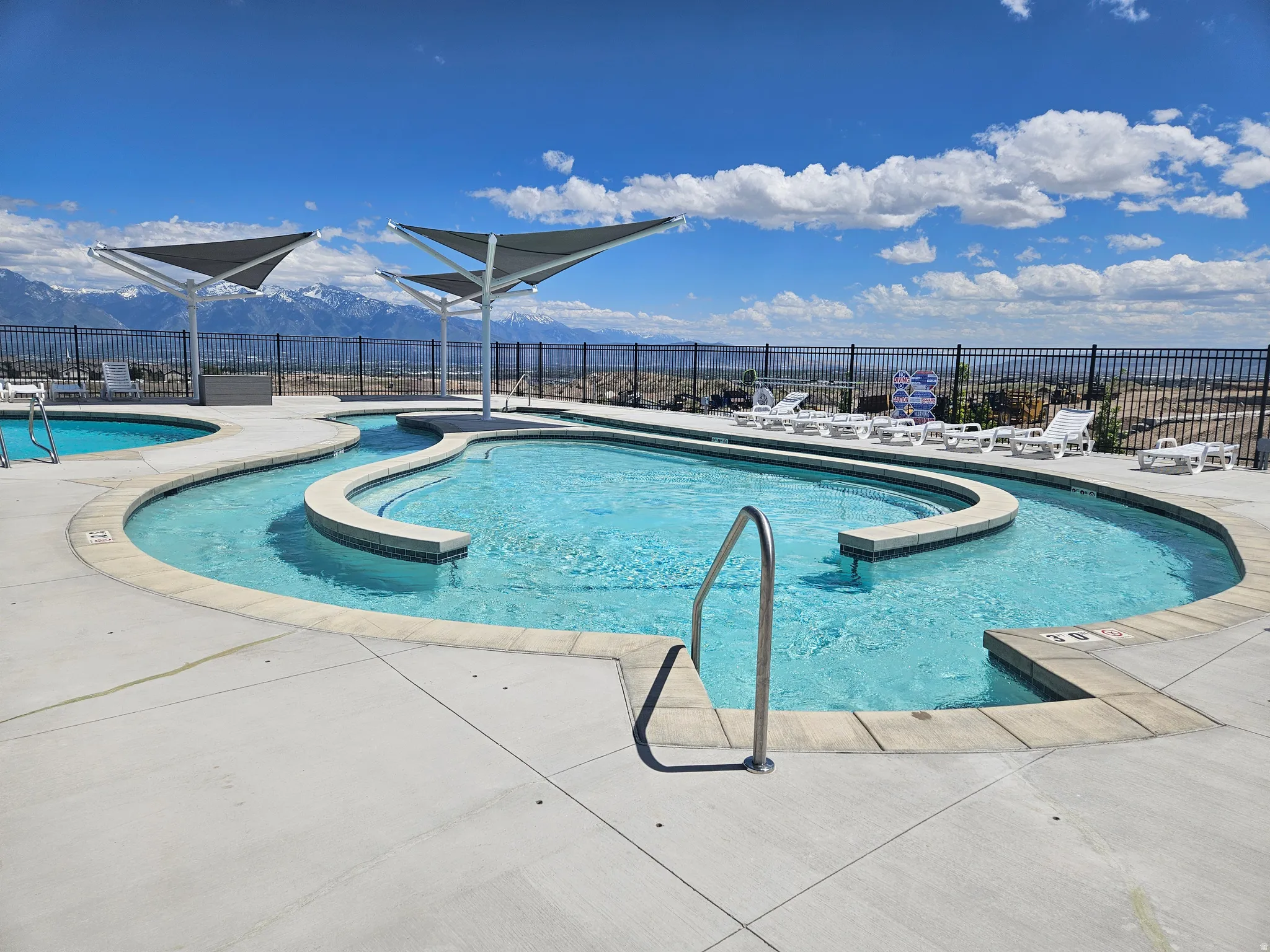 Curved swimming pool with light blue water, surrounded by concrete decking and a black metal fence