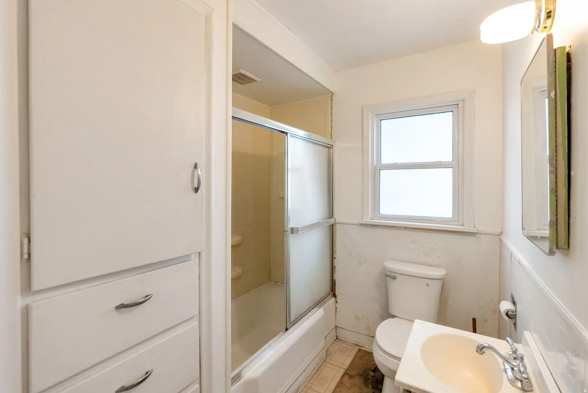 Bathroom featuring a built-in storage cabinet with drawers, a tub-shower combination with sliding glass doors, a window, a toilet, and a vanity with a white sink