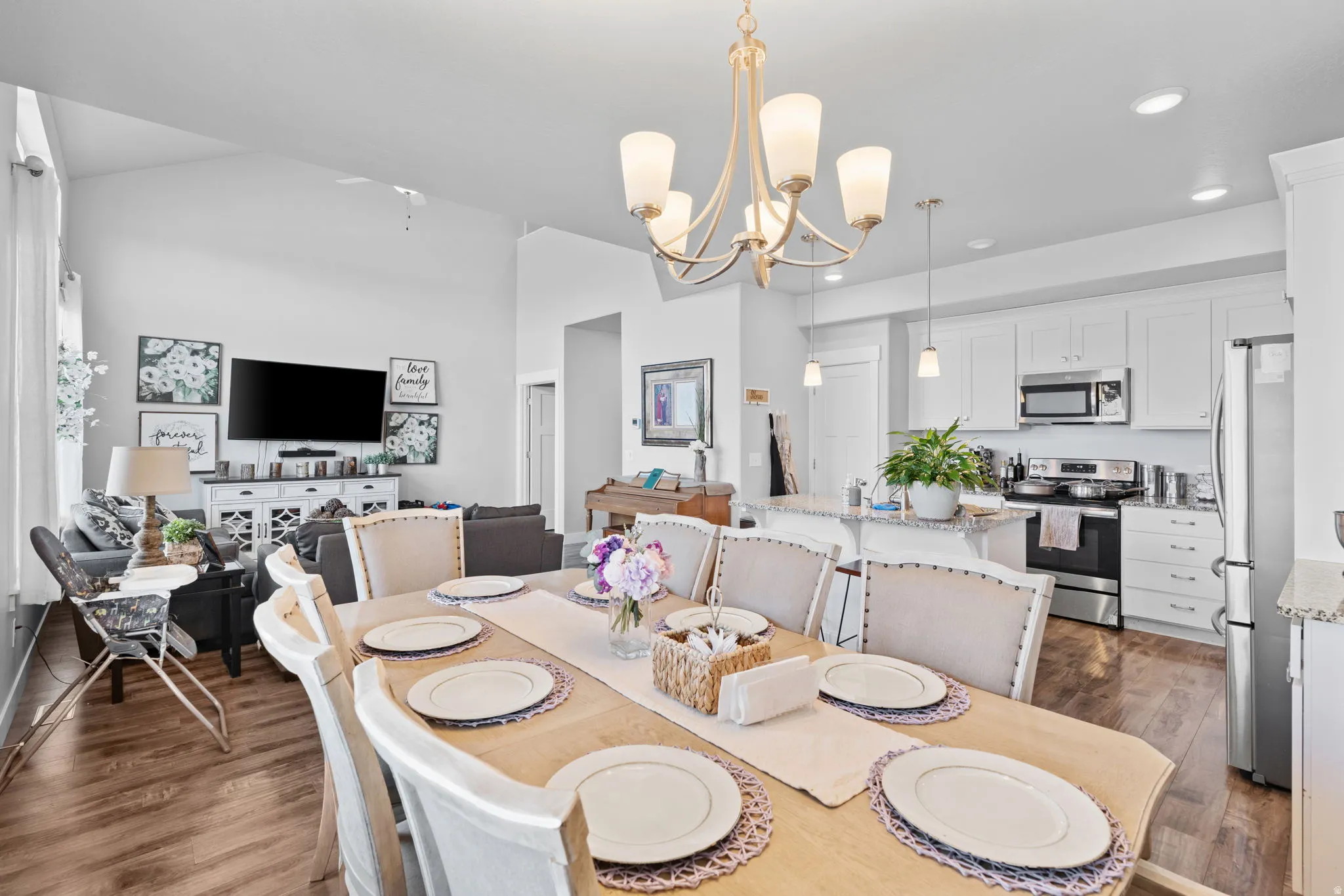 Open-concept living space featuring wood-finish flooring, a contemporary kitchen with white cabinetry and stainless steel appliances, a breakfast bar, recessed lighting, and a dining area illuminated by a decorative chandelier