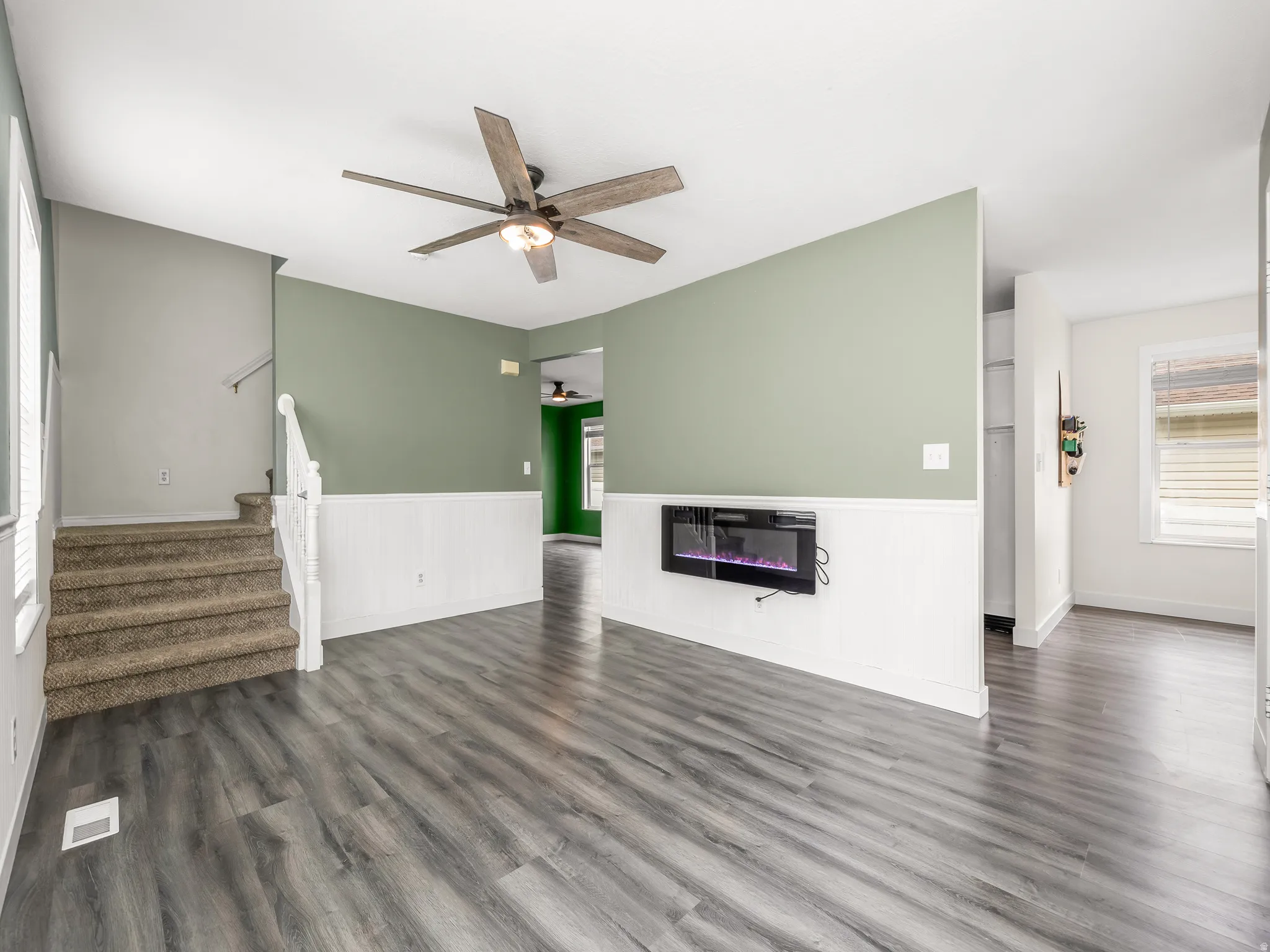 Spacious interior featuring wood-finish flooring, a wall-mounted electric fireplace, white wainscoting, and a ceiling fan with integrated lighting