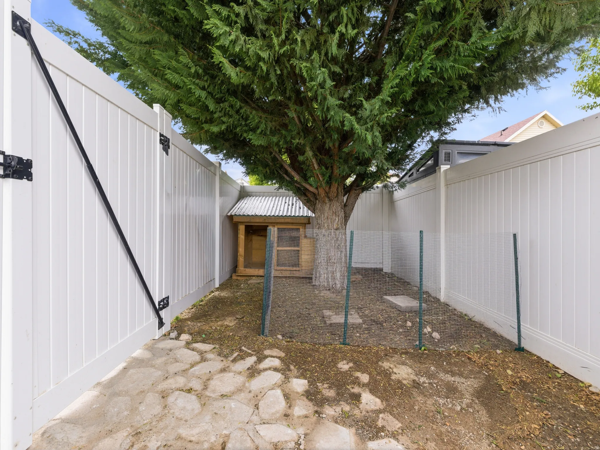 Fenced outdoor area featuring chicken coop, a mature tree, stone pathway, and a wood-structure shed with a corrugated roof