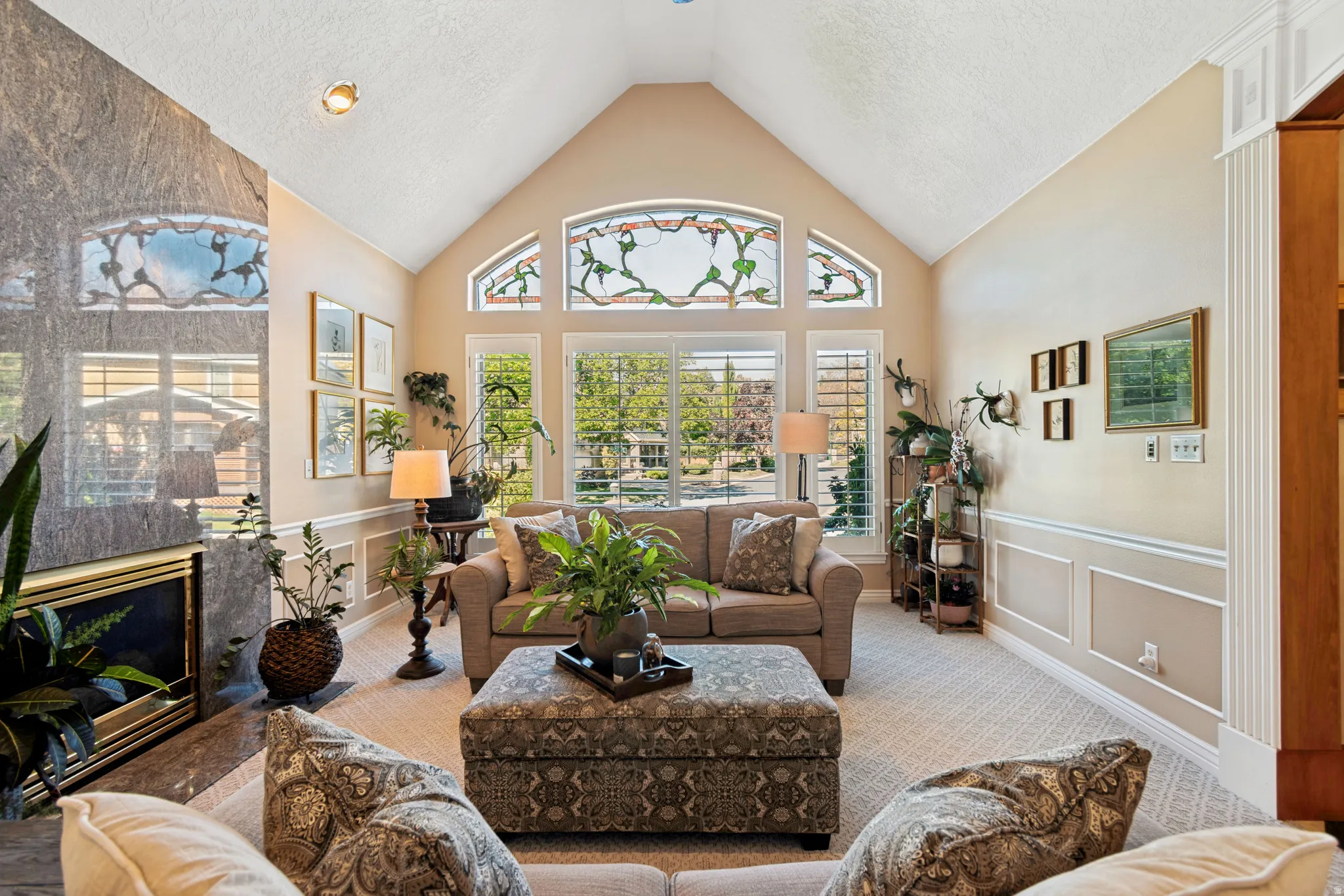 Vaulted ceiling with decorative stained glass windows, large windows with plantation shutters, a fireplace with a stone surround, and wainscoting