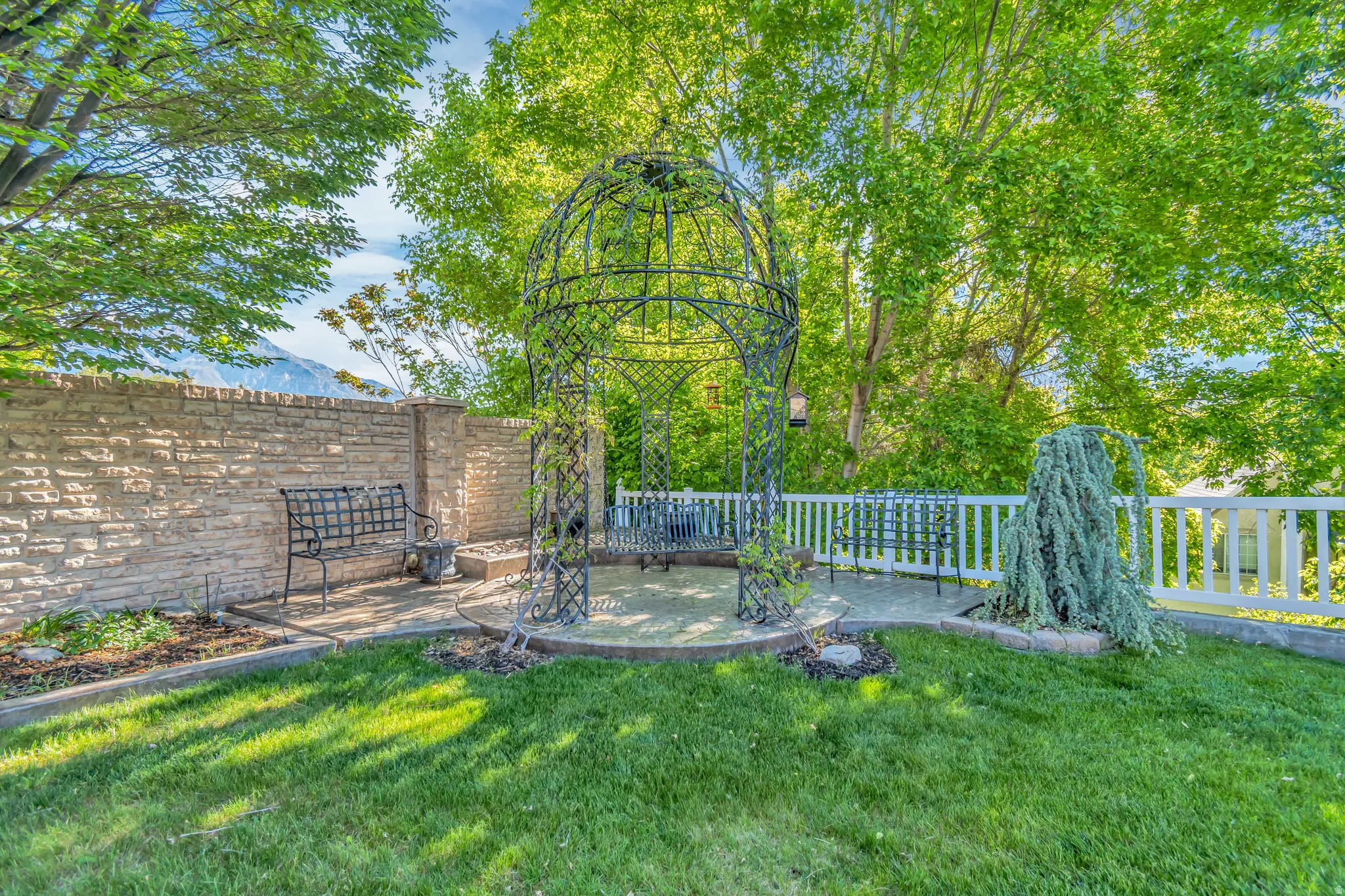 Expansive backyard featuring a wrought iron gazebo, stone retaining wall, and a white picket fence