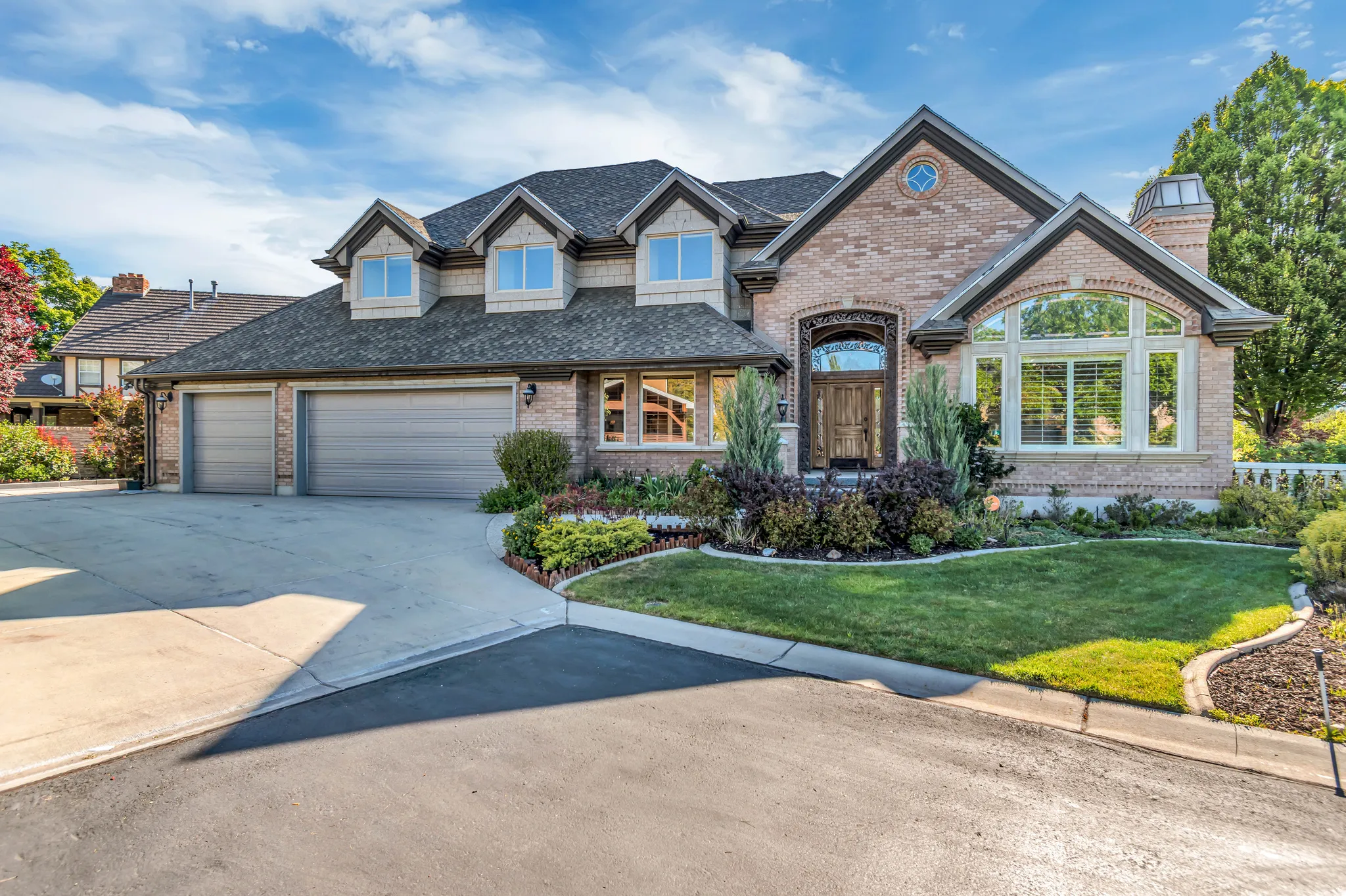 Brick exterior featuring a multi-car garage, mature landscaping, arched entry, and prominent bay window