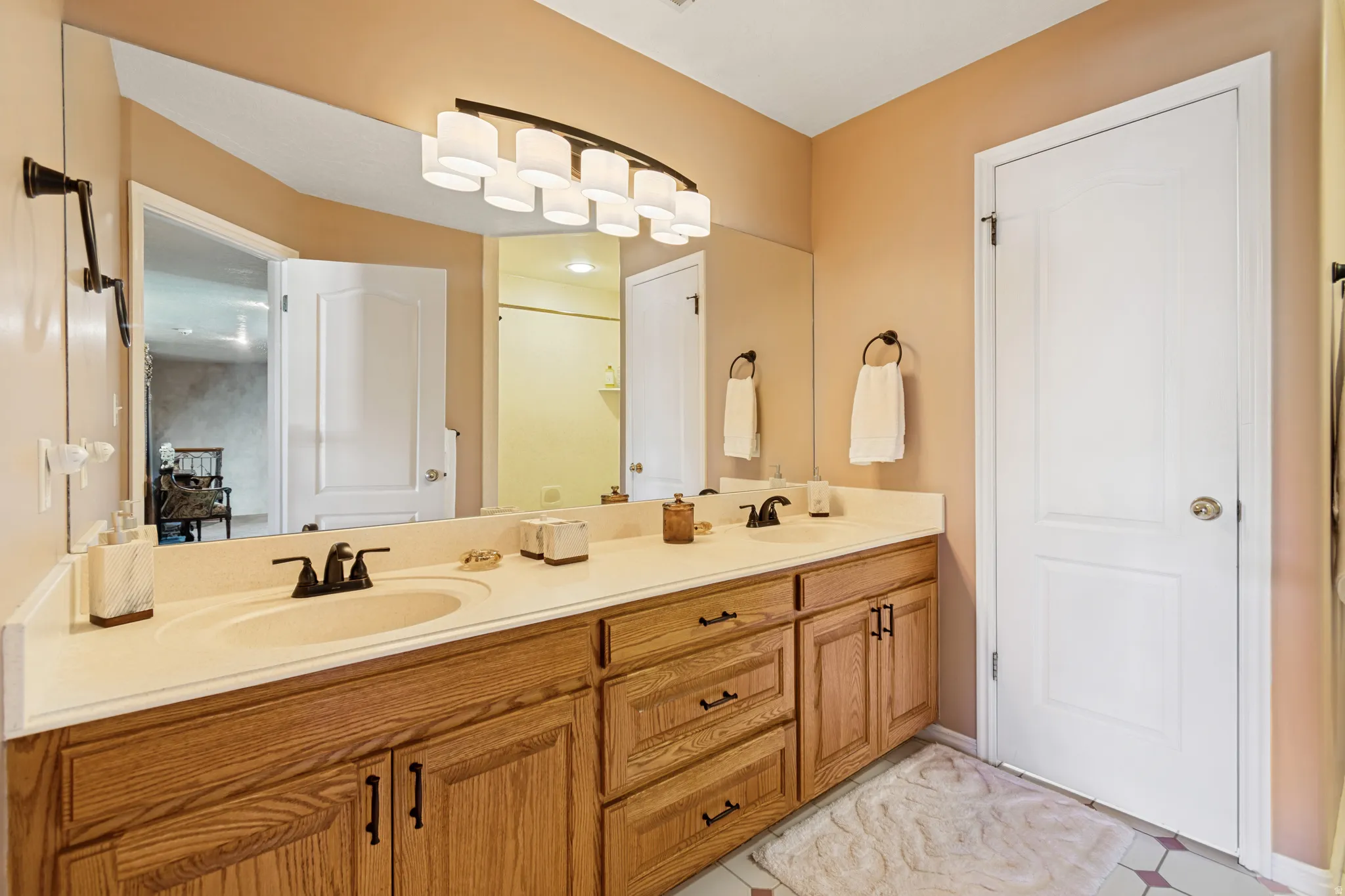 Bathroom featuring a dual vanity with light wood-finish cabinetry, a large wall-mounted mirror, and a multi-light fixture with white shades