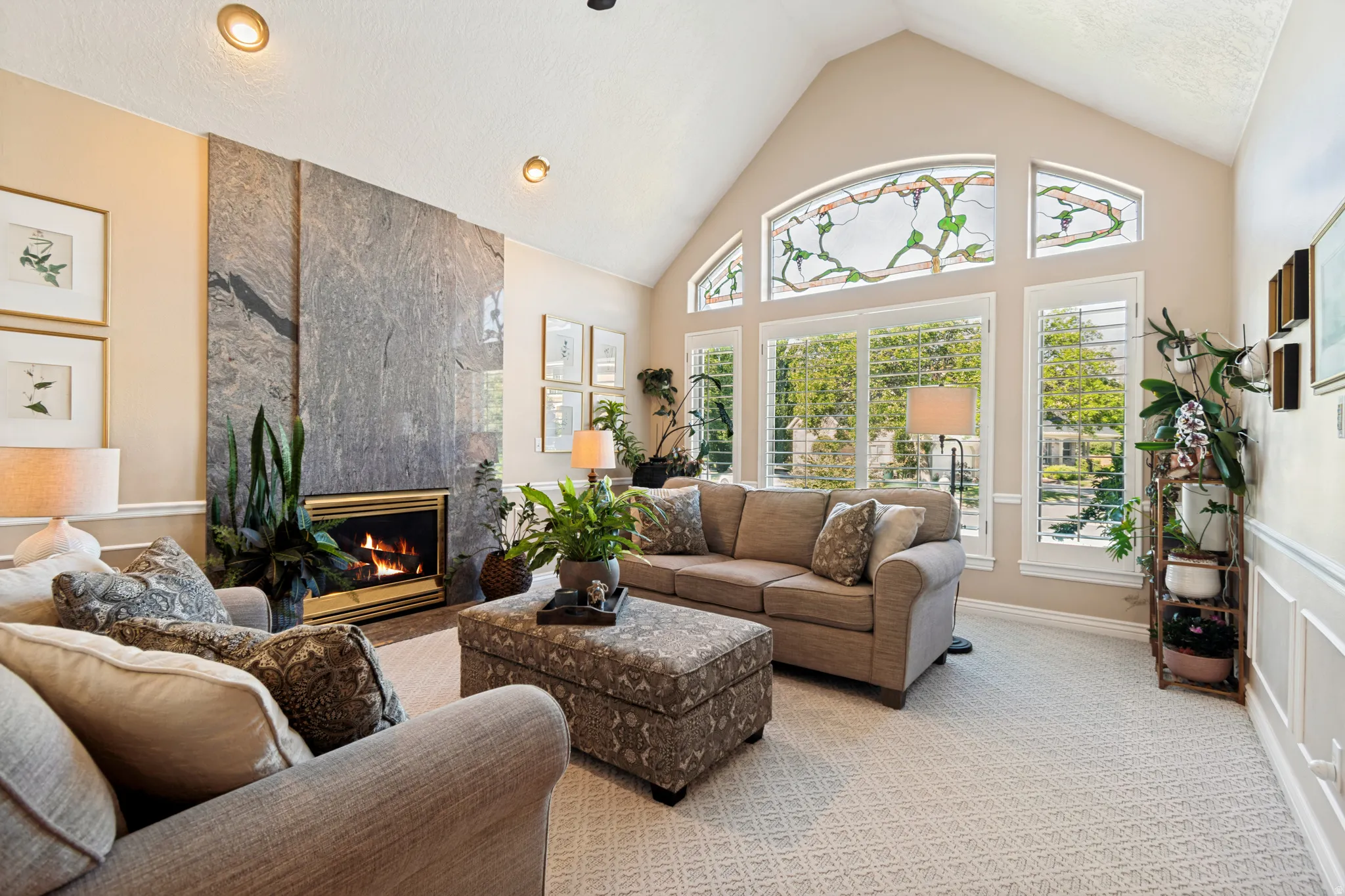 Vaulted ceiling living area featuring a full-height stone fireplace surround, multiple transom windows with stained glass elements, and plantation shutters