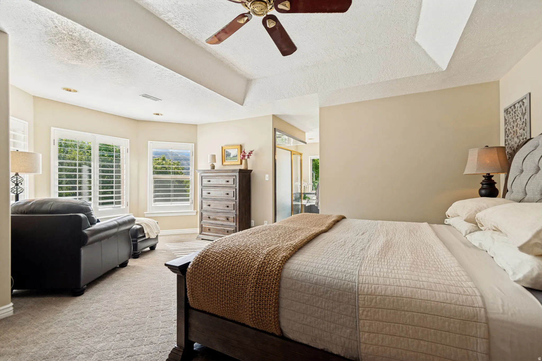 Spacious bedroom featuring a tray ceiling with a ceiling fan, bay windows with plantation shutters, plush carpet flooring, and neutral wall tones