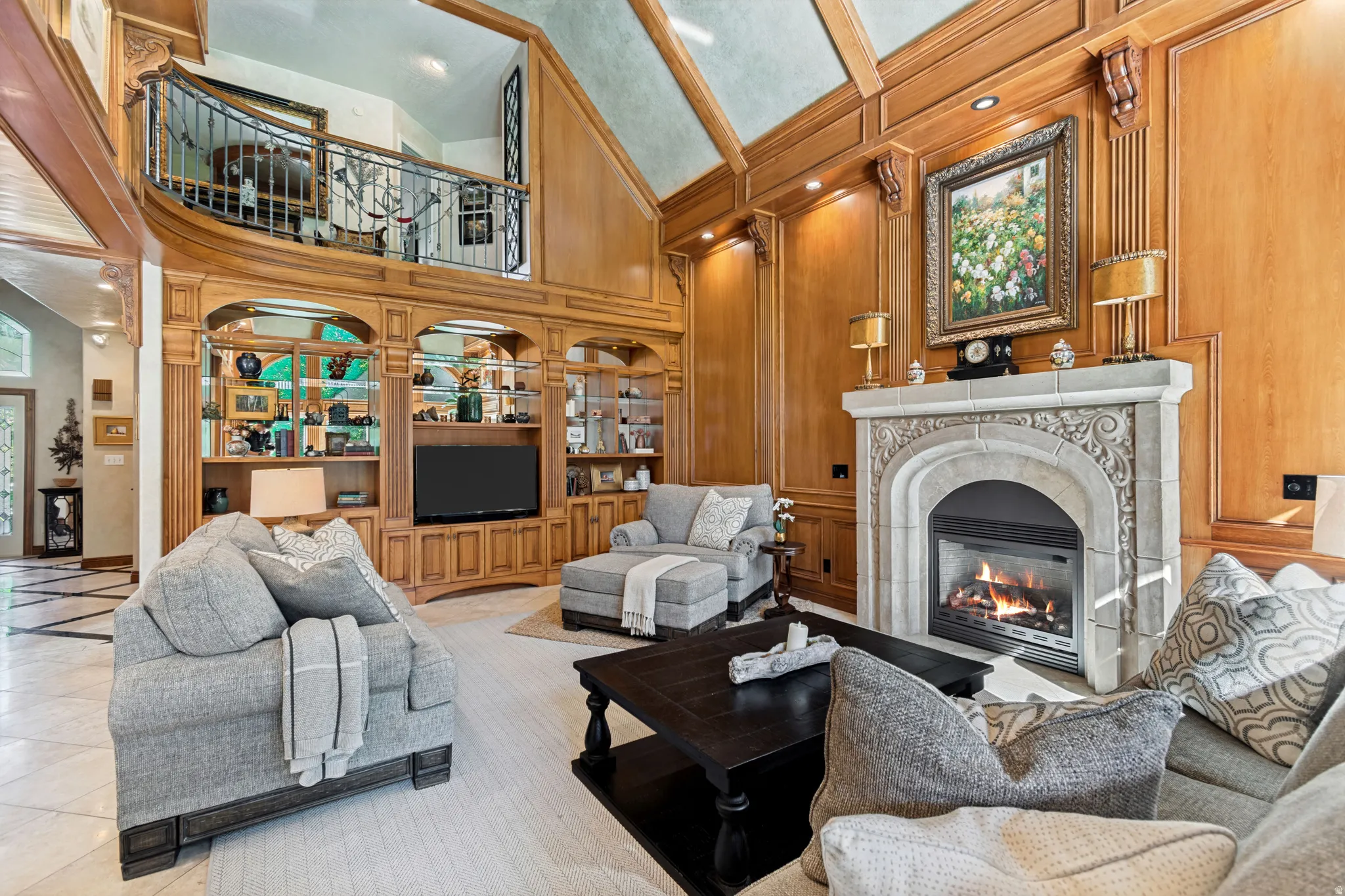Living area featuring a grand stone fireplace with intricate carvings, extensive wood paneling, a built-in entertainment center with display shelving, and an overhead balcony with iron railings