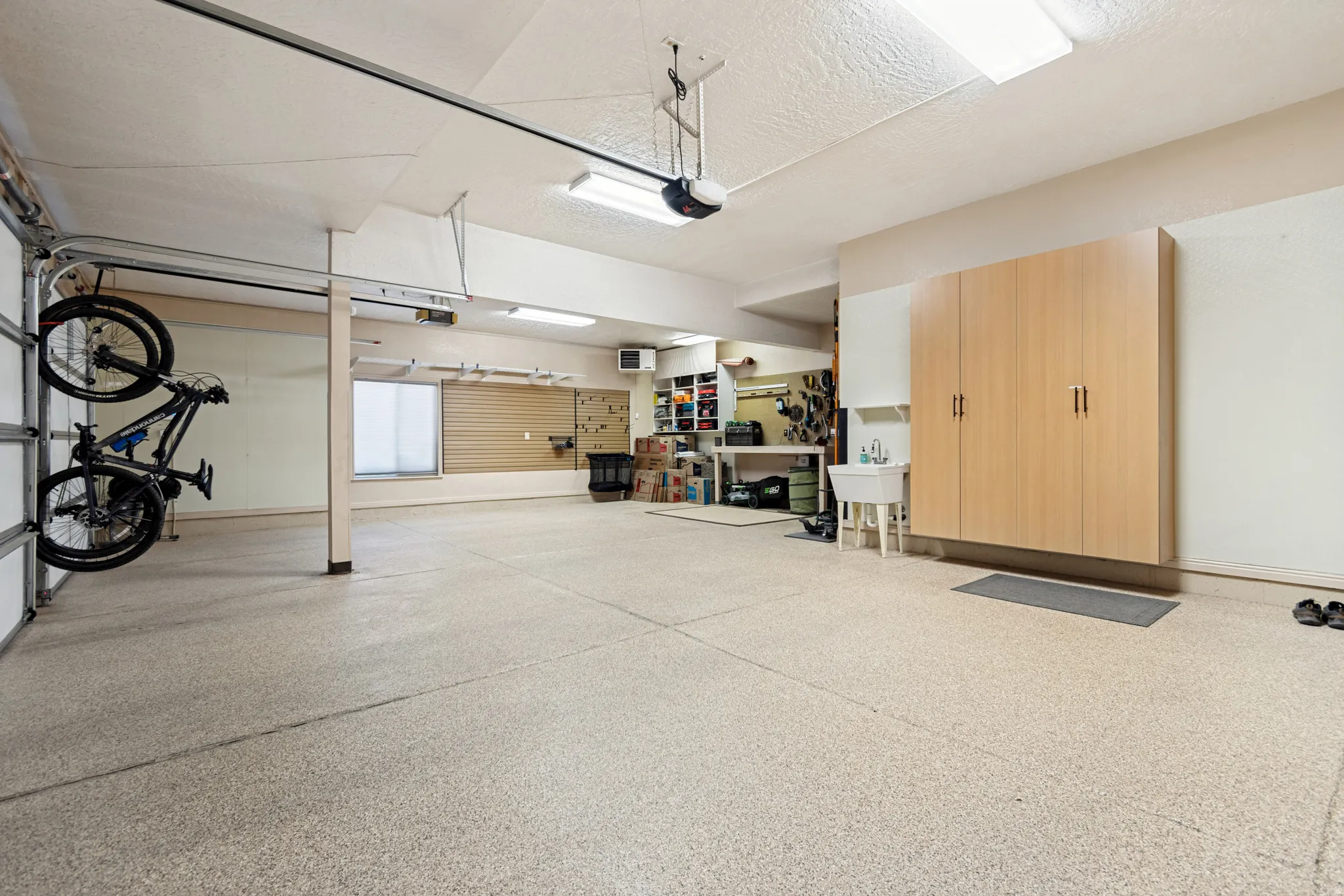 Spacious garage featuring a light-colored epoxy floor, ample overhead lighting, built-in wood-finish cabinetry, and a utility sink