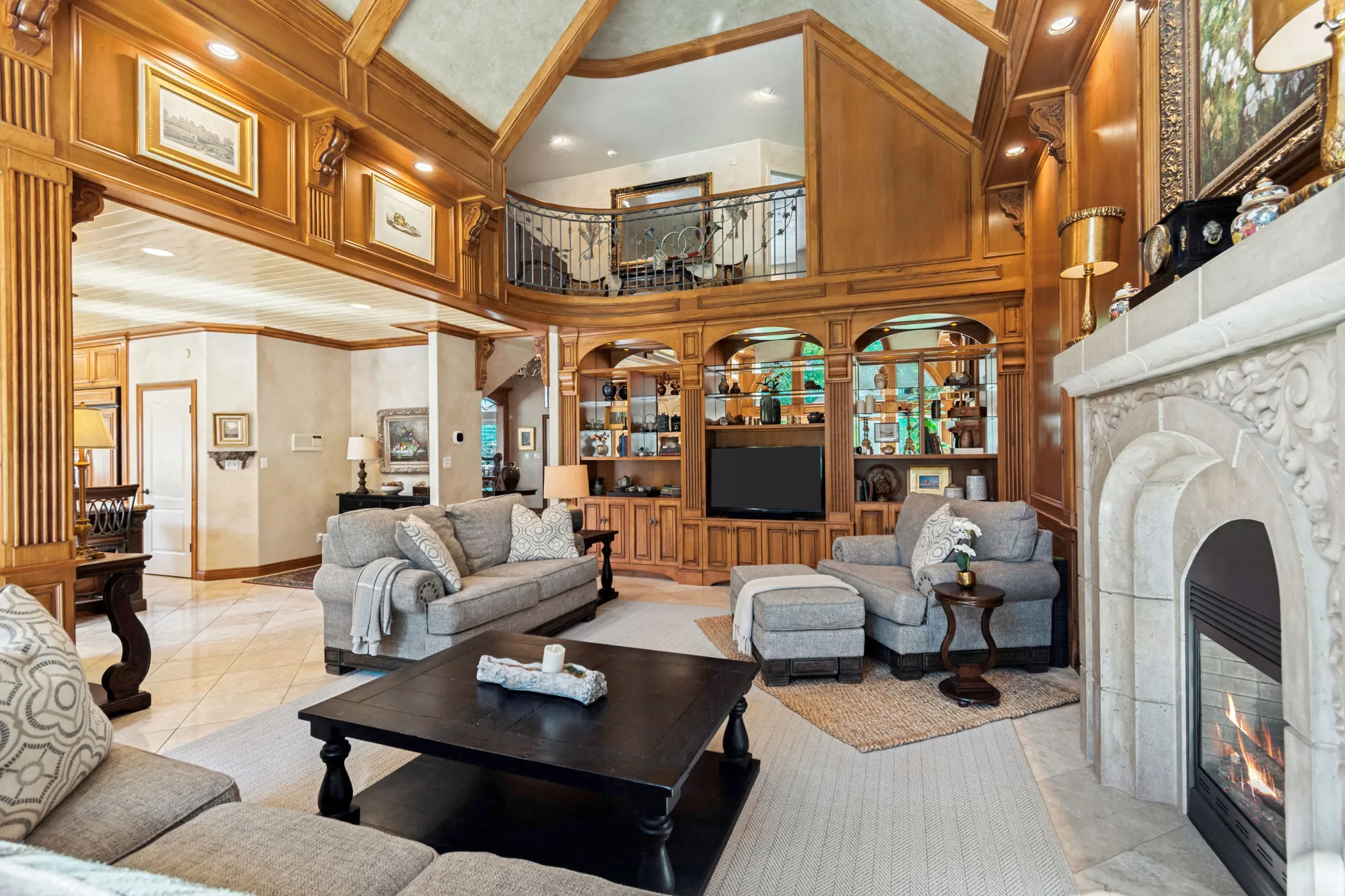 Spacious living area featuring a stone-carved fireplace, custom built-in cabinetry, wood-paneled walls, a second-story balcony with wrought iron railing, and a vaulted ceiling with exposed wood beams