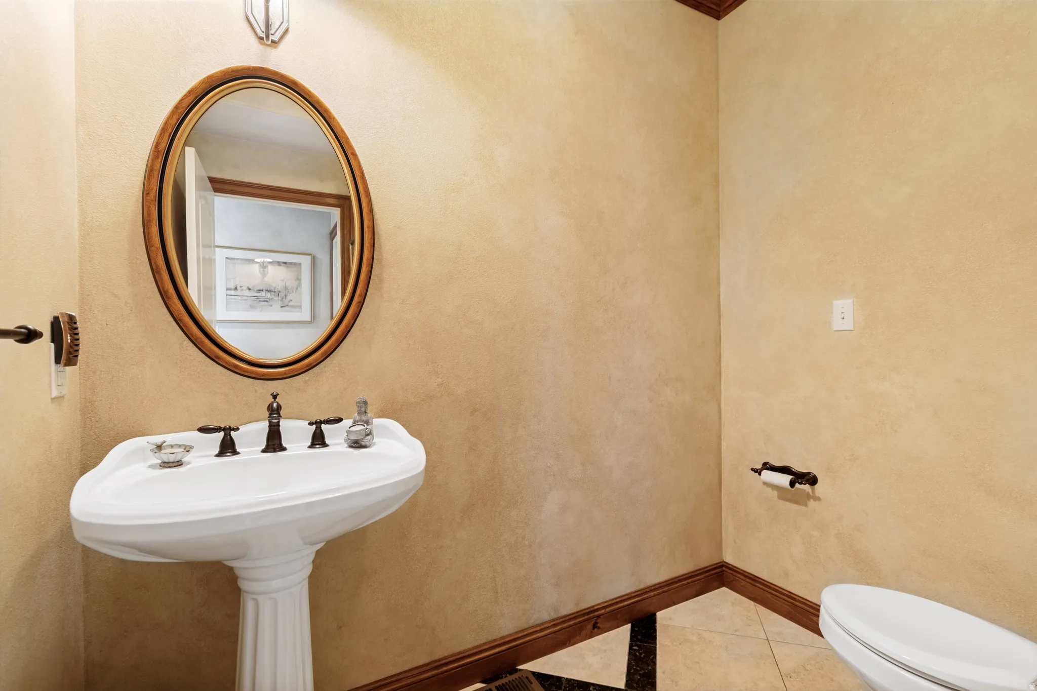 Pedestal sink with bronze-finish fixtures and an oval wood-framed mirror