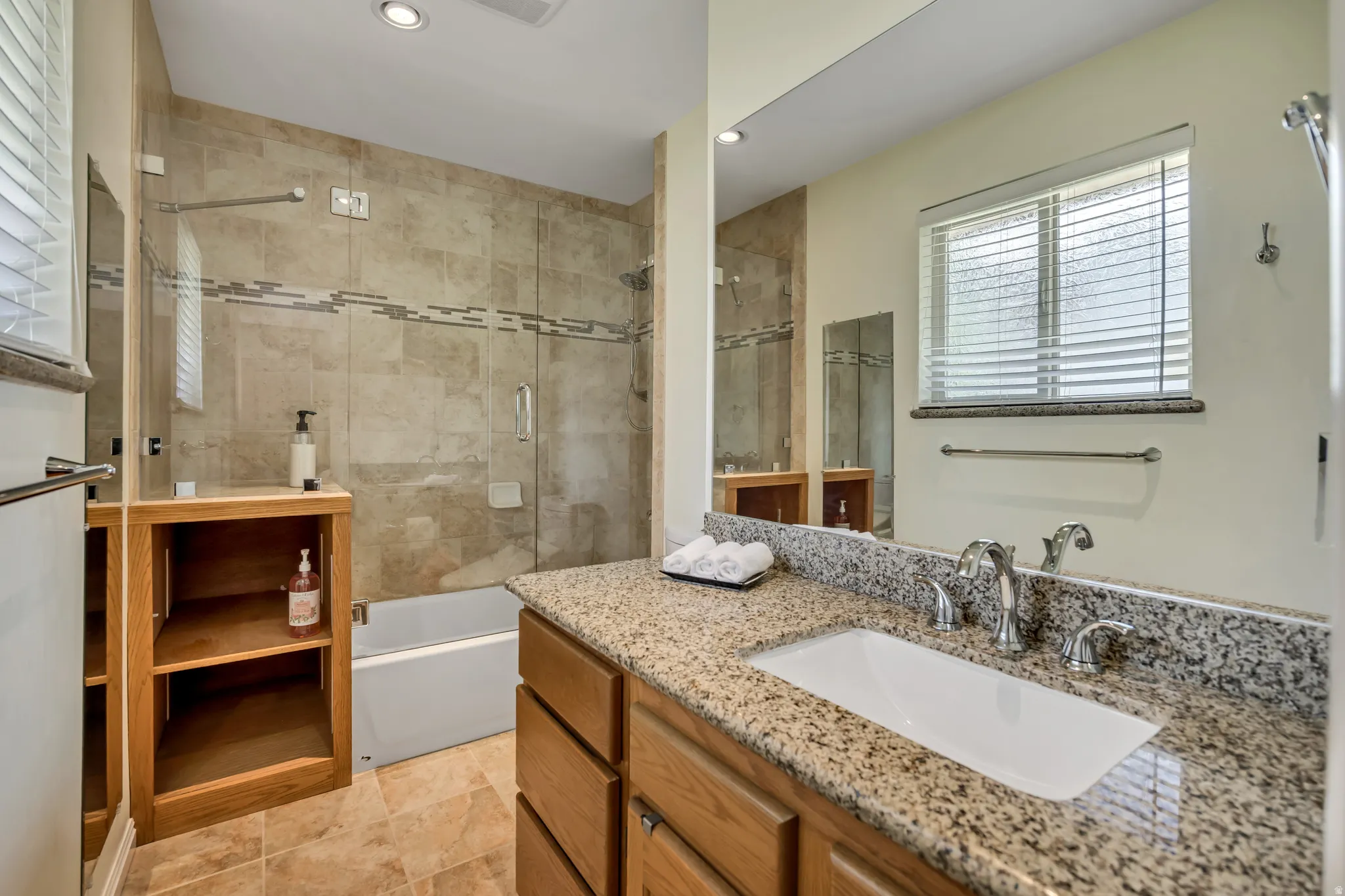 Bathroom featuring a tiled shower/tub combination with glass enclosure, a vanity with a granite-style countertop and undermount sink, a recessed wall mirror, a window with blinds, and tiled flooring
