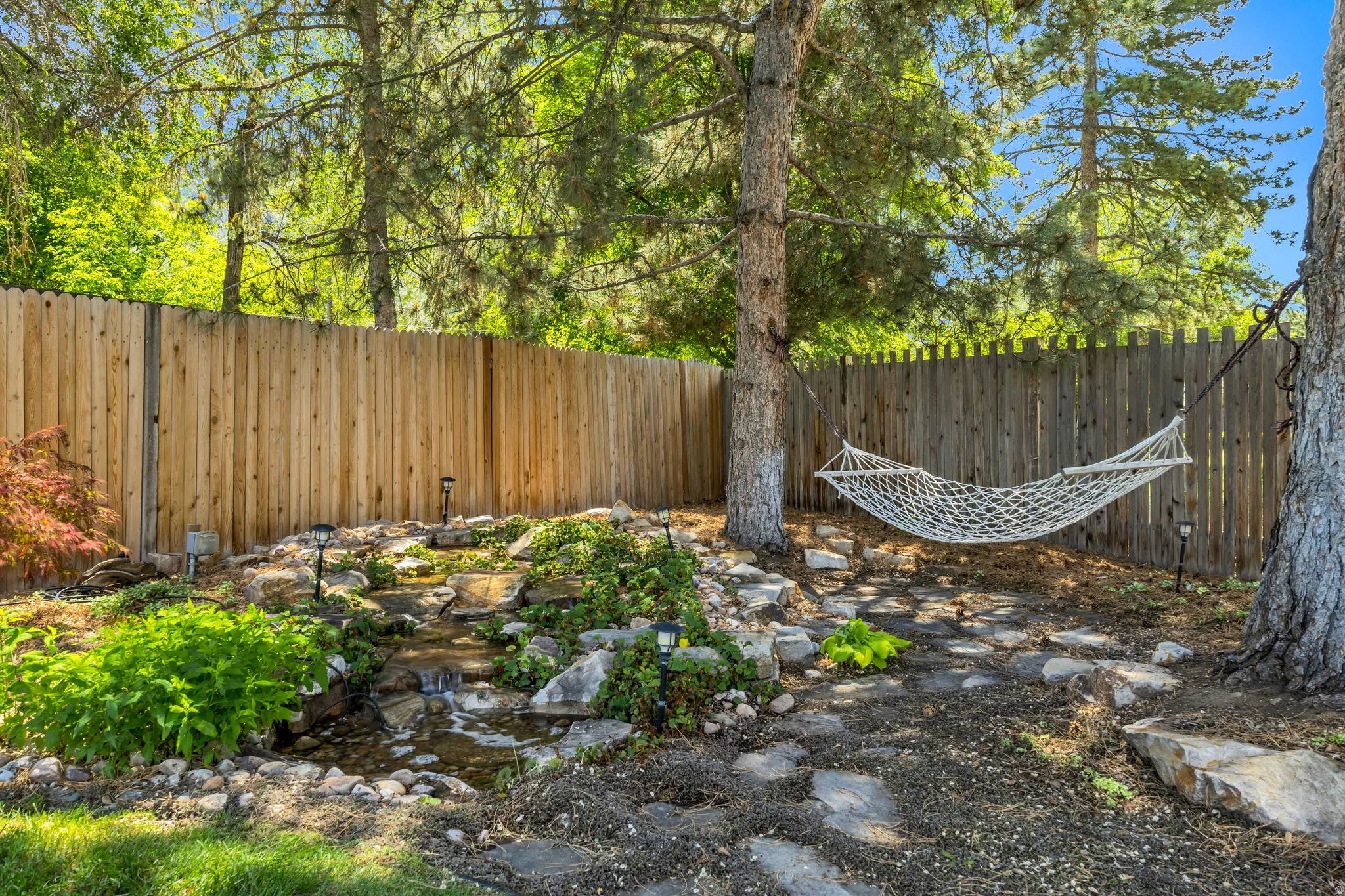 Backyard sanctuary featuring a multi-tiered water feature with rock accents, mature trees, a wooden privacy fence, and stepping stone pathways