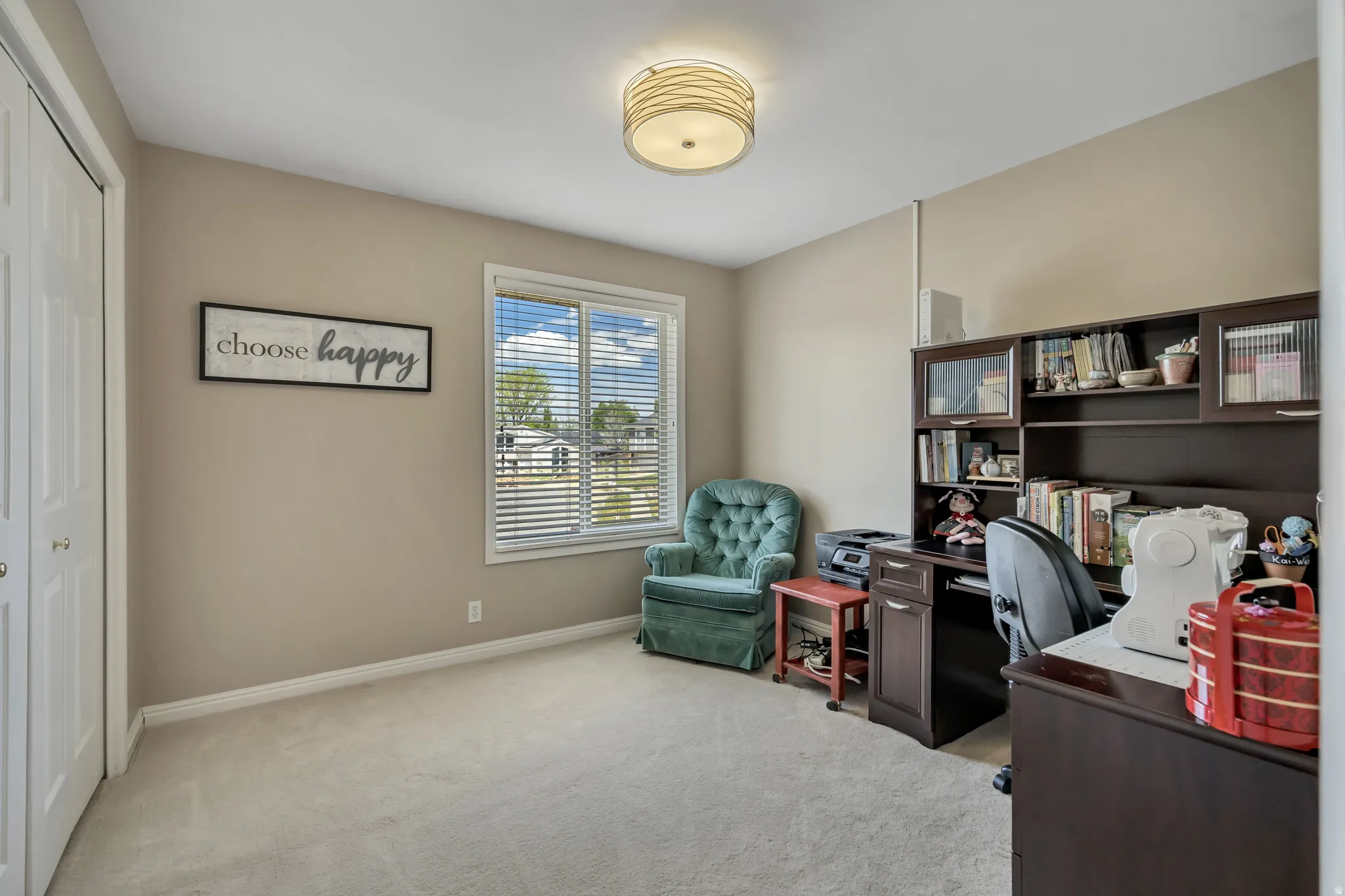 Versatile room featuring a window with horizontal blinds, built-in desk with shelving, light-colored carpeting, and a ceiling-mounted drum light fixture
