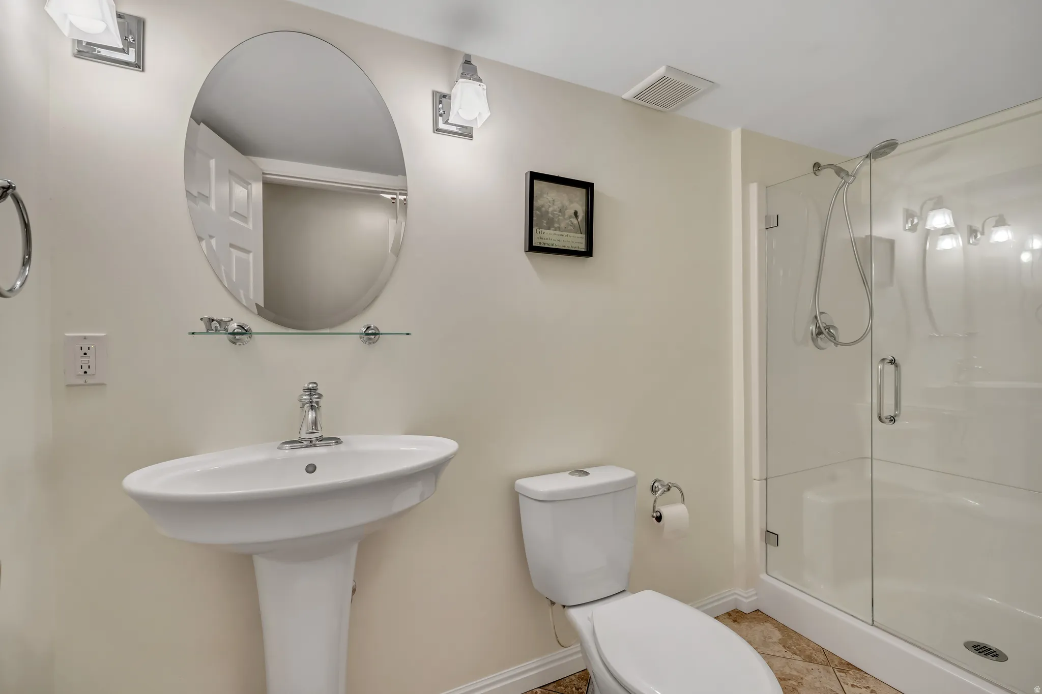 Bathroom featuring a pedestal sink, oval mirror with sconce lighting, and a glass-enclosed shower with a handheld showerhead
