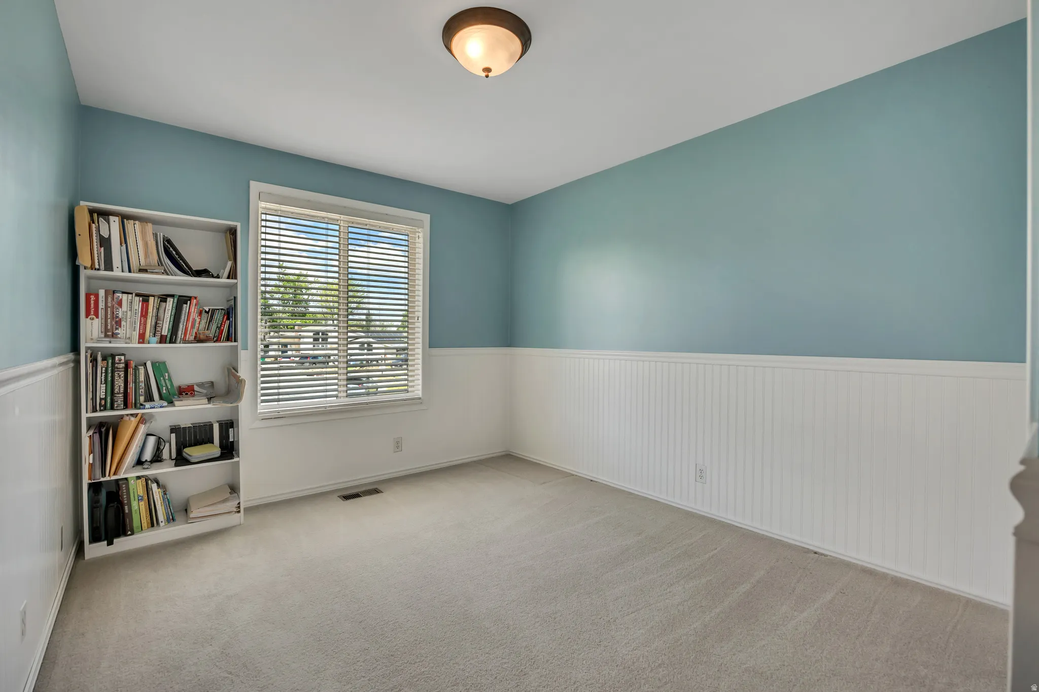 Light grey carpeted room featuring a window with blinds, a white-paneled wainscoting, and a semi-flush ceiling light fixture