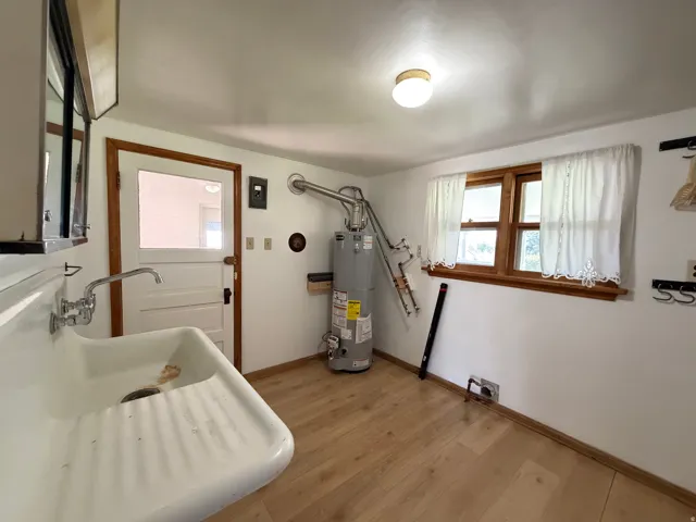 Utility room featuring a utility sink, wood-finish flooring, a window with wood trim, and a water heater