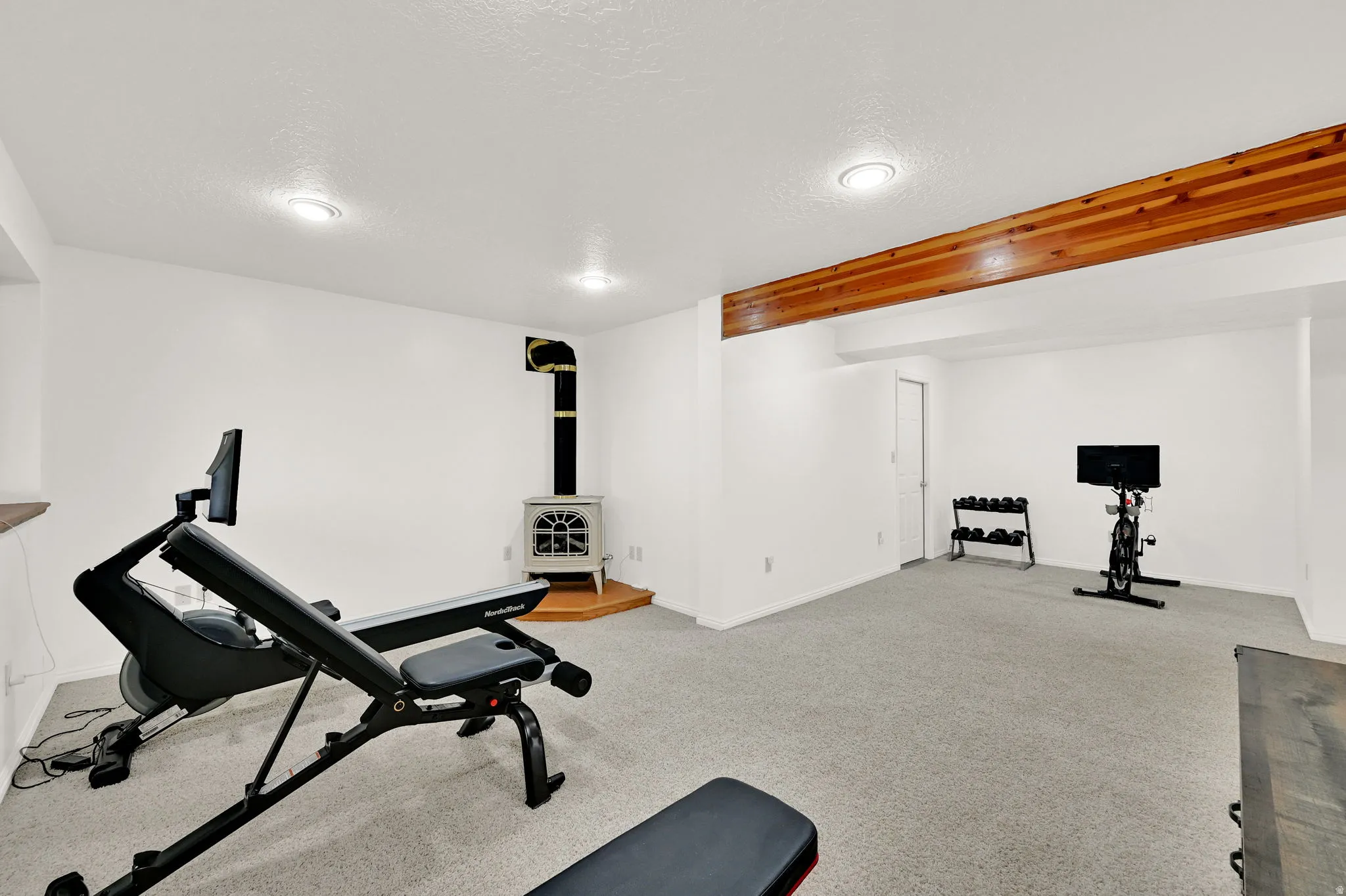 Spacious room featuring light gray carpeting, crisp white walls, recessed ceiling lighting, a prominent exposed wood beam, and a white freestanding stove with a black stovepipe