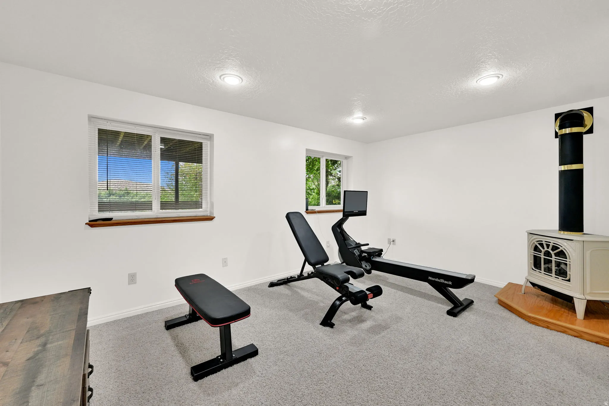 Spacious room featuring light gray carpeting, recessed ceiling lighting, a cream-colored freestanding stove with a black stovepipe, and white walls