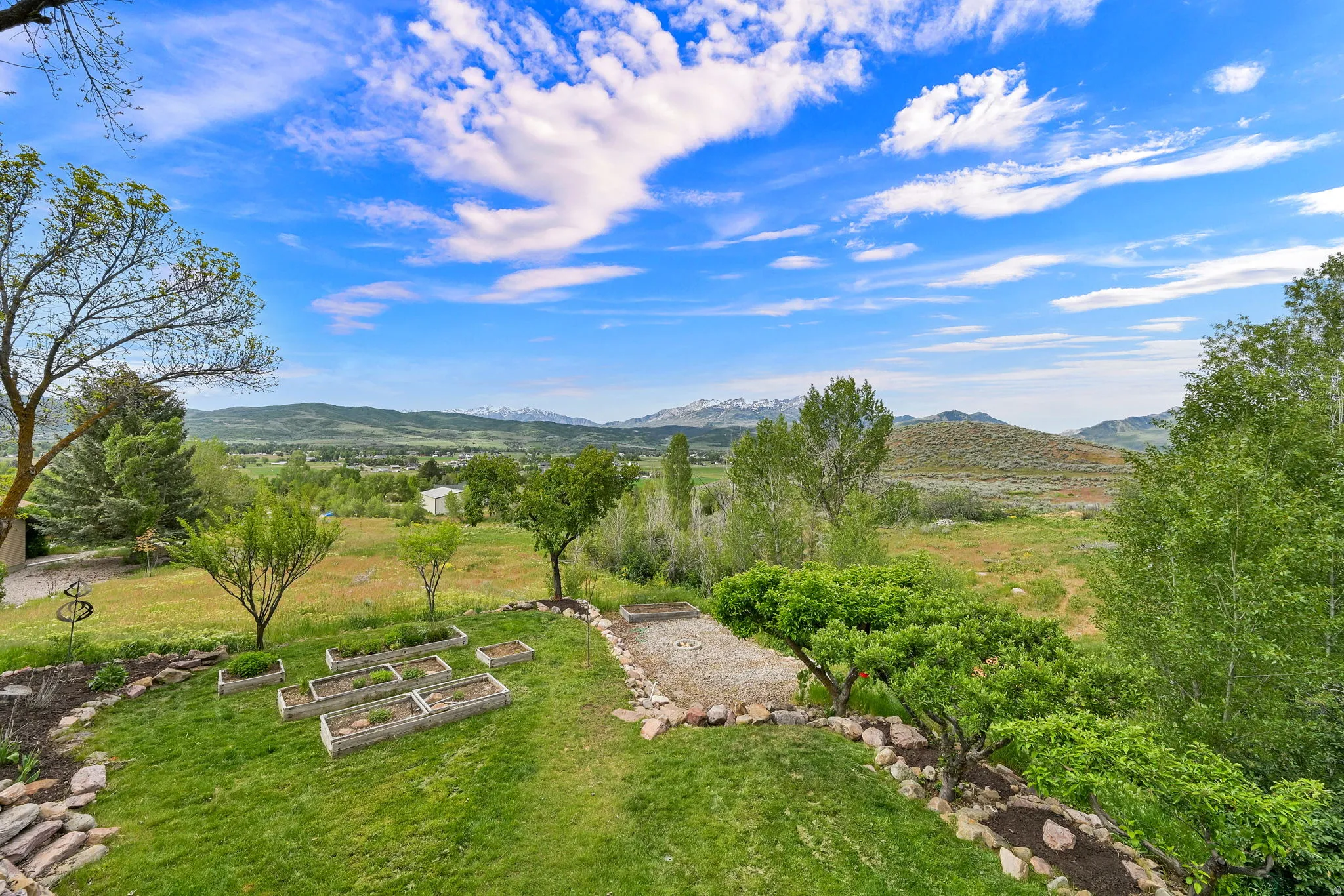 Expansive backyard featuring a manicured lawn, multiple raised garden beds, established trees, and a rock-lined hardscaped area