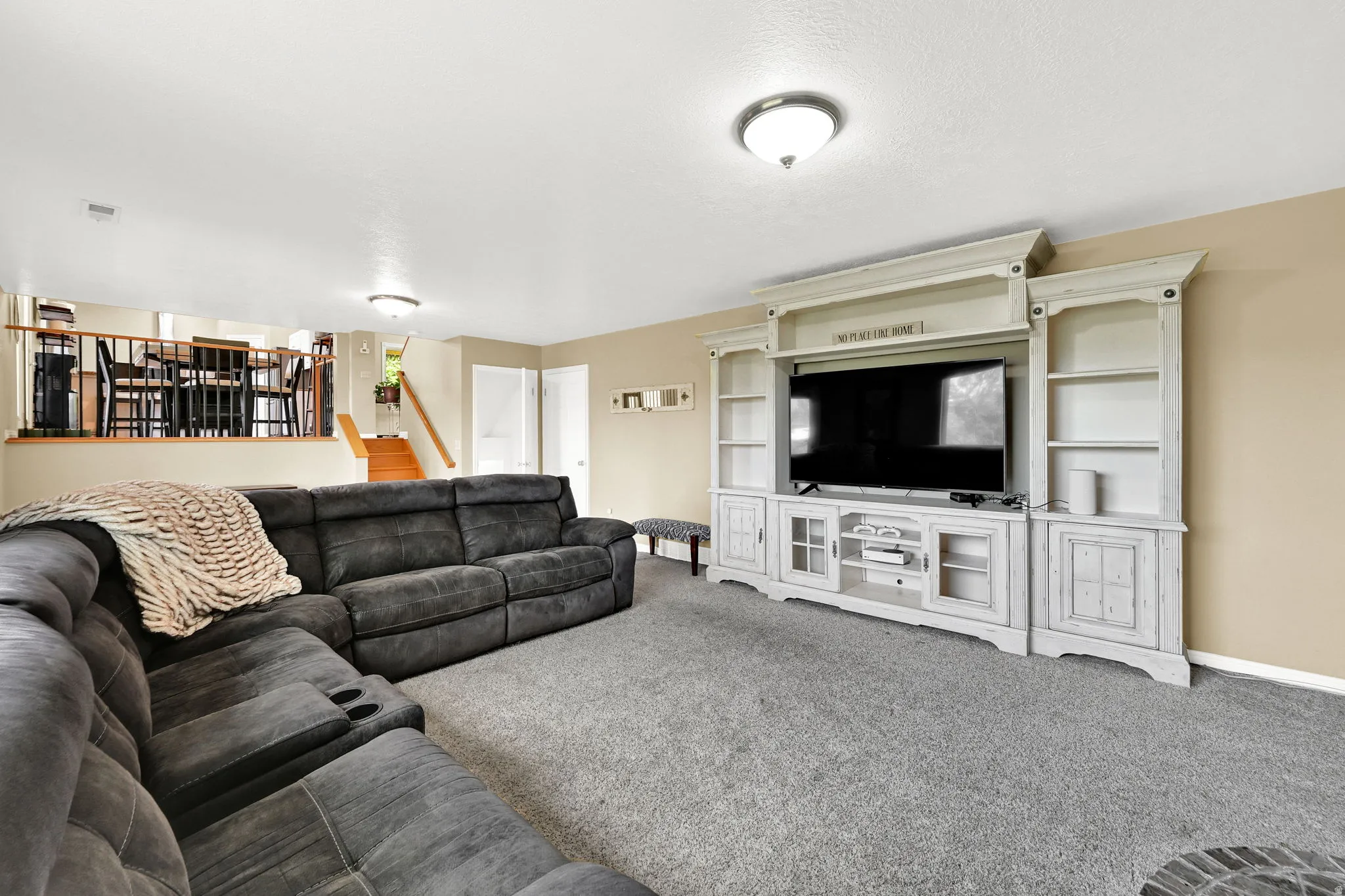 Carpeted living area featuring a built-in media console with shelving and cabinet storage, ceiling-mounted lighting, and a partial view of a staircase with wood treads and a railing