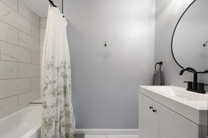 Bathroom featuring a white bathtub with subway tile surround, white vanity, black hardware, and a circular mirror