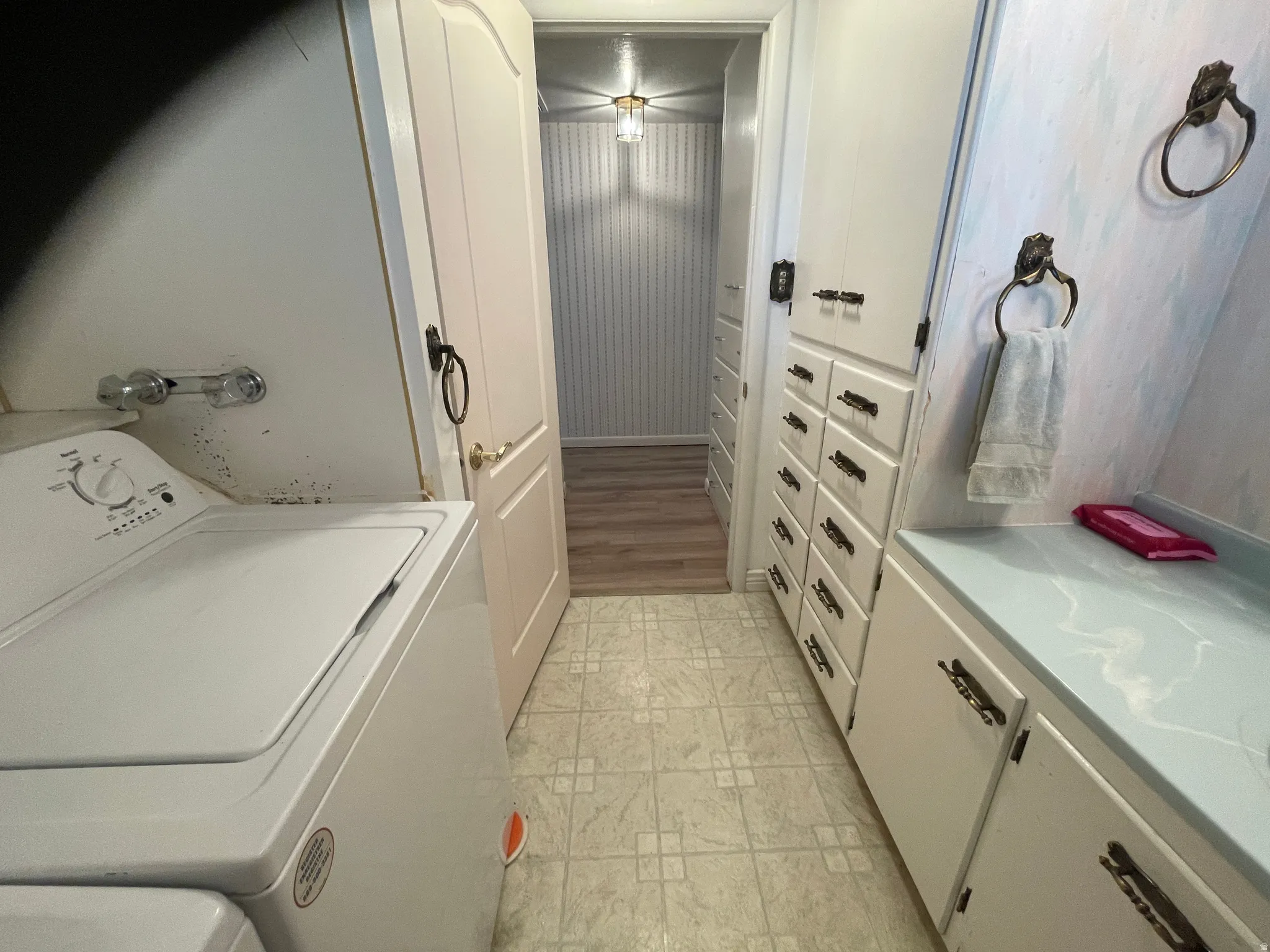 Compact laundry and half bath, featuring a tile floor, white cabinetry, a light blue countertop, and a hallway beyond with wood-finish flooring