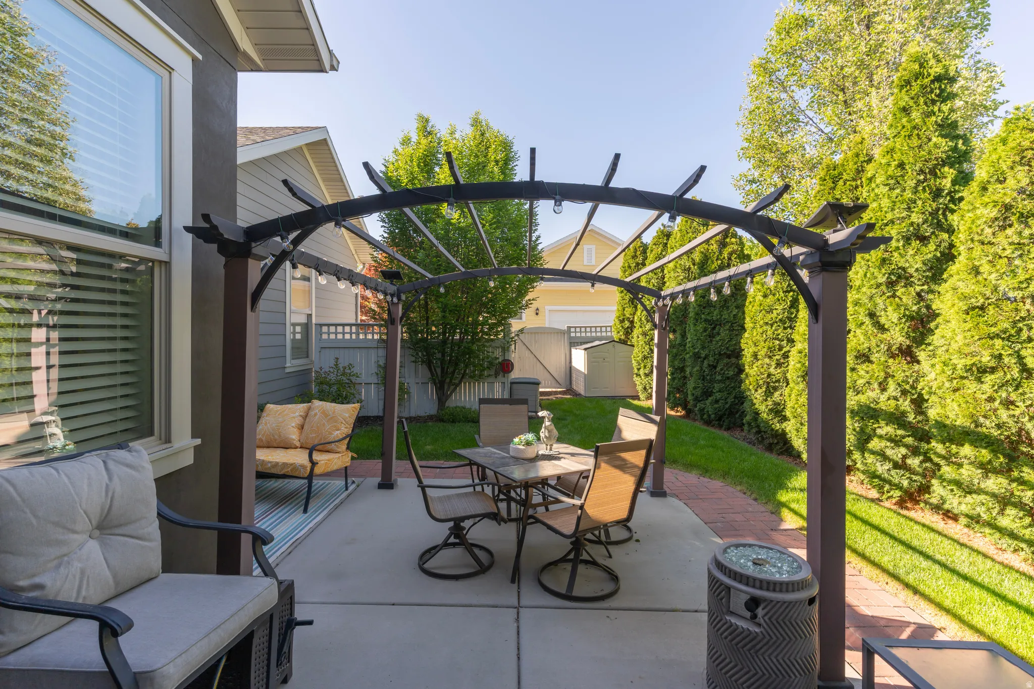 Outdoor patio featuring a pergola with string lighting, concrete slab, brick pathway, and a manicured lawn with mature privacy trees