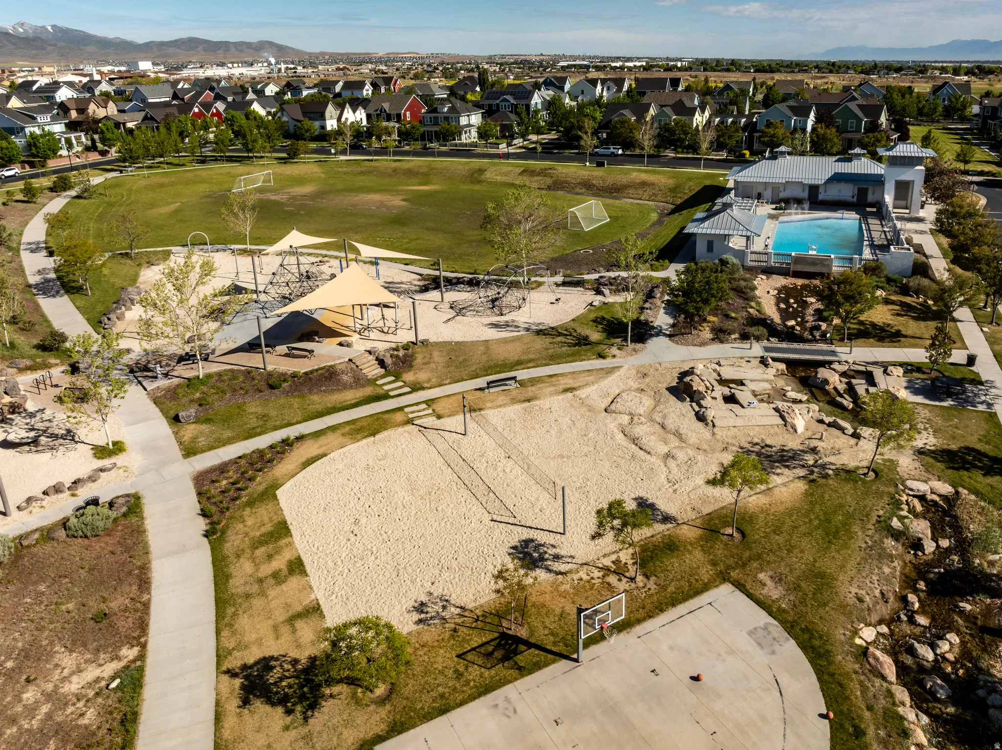 Expansive community park featuring a swimming pool, shaded playground area, sandy volleyball court, basketball half-court, and open green spaces