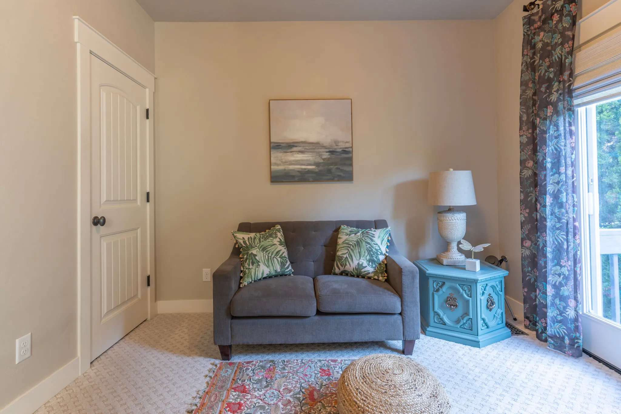Interior room featuring neutral wall tones, a white paneled door with dark hardware, light patterned carpet flooring, white baseboards, and a glass-paneled exterior door with floral drapes