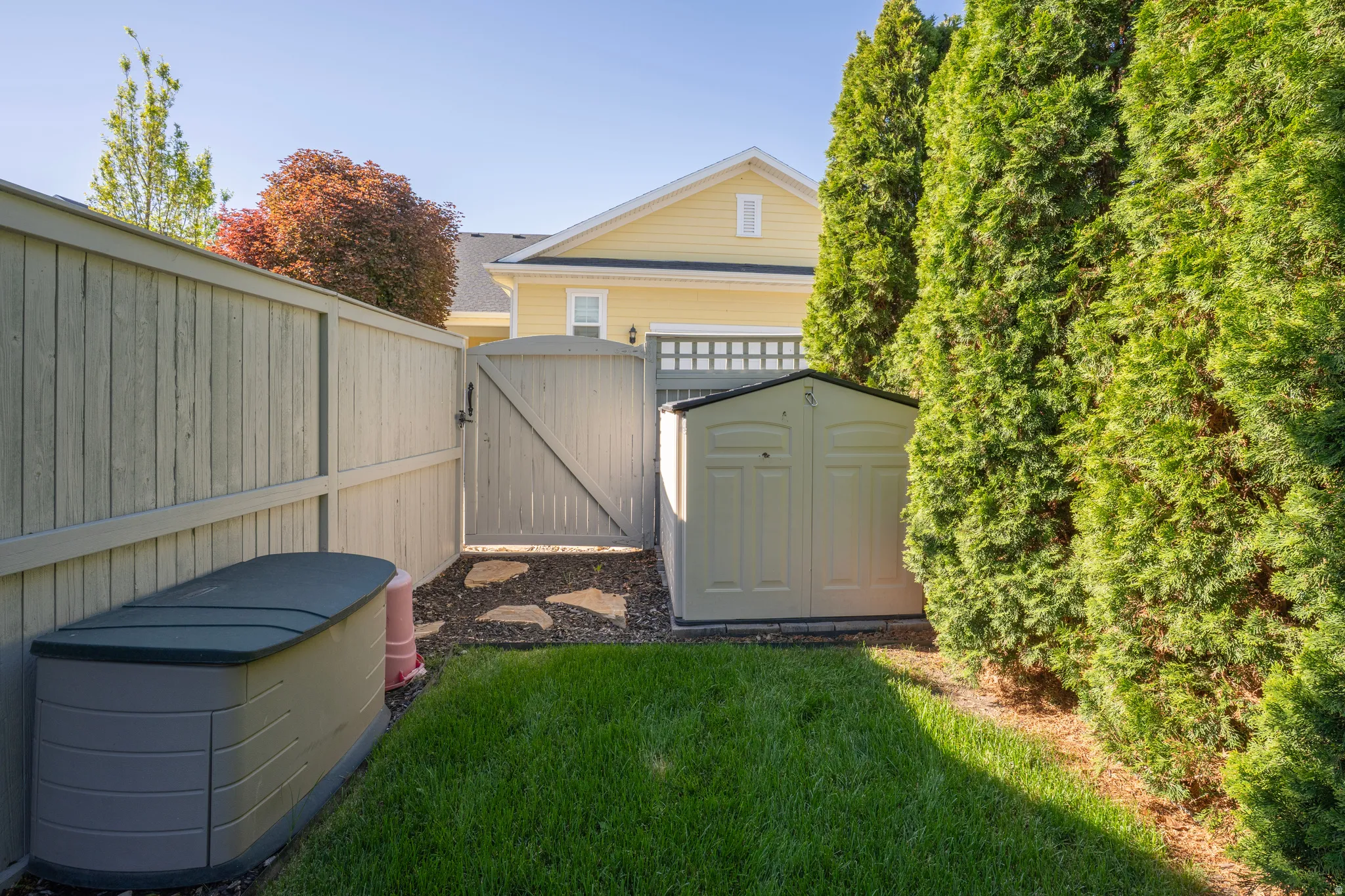 Fenced side yard featuring a grass lawn, privacy gate, evergreen shrubs, and two storage sheds