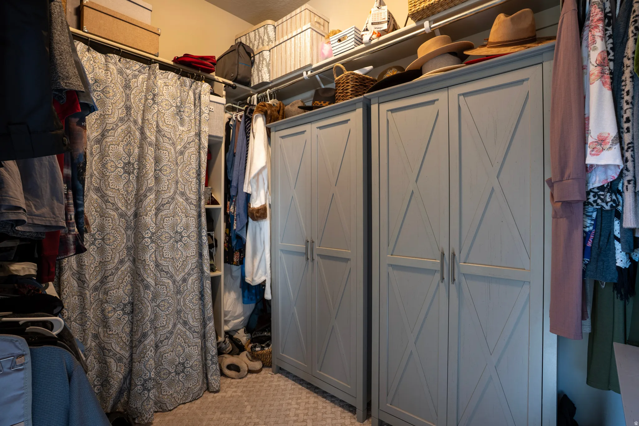 Custom closet system featuring light gray paneled cabinetry with X-detail, a patterned fabric curtain, built-in shelving, and carpet flooring