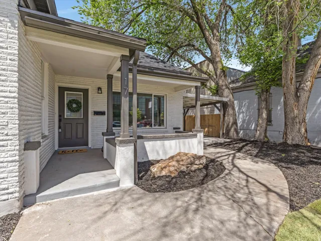 Covered front porch featuring a dark gray door with glass insert, light brick exterior, dark gray trim, and mature trees providing natural shade