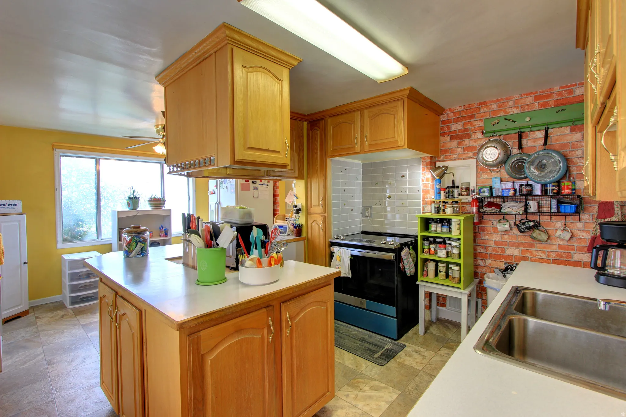 Spacious kitchen featuring an island with built-in cabinetry, dual basin stainless steel sink, wood-finish cabinetry, and brick-patterned accent wall