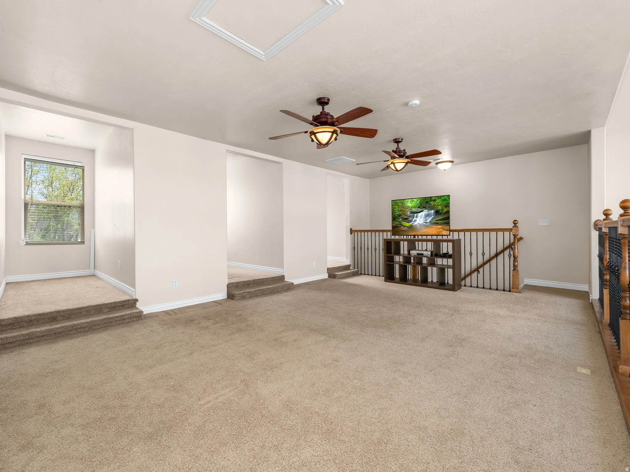 Carpeted loft area featuring multiple ceiling fans with integrated lighting, a railing with wood newel posts and wrought iron spindles, a window with horizontal blinds, and a stepped platform
