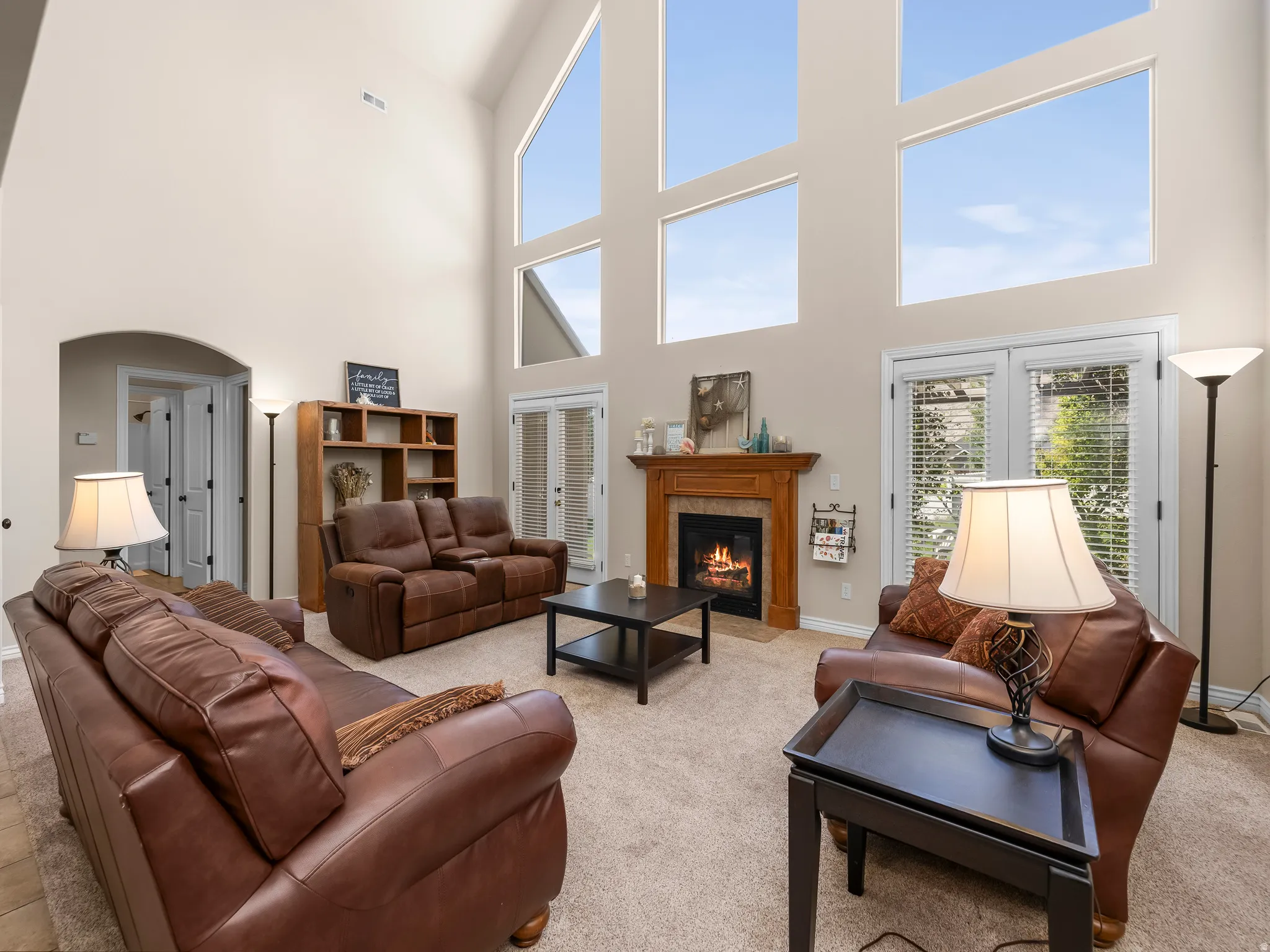 Living area featuring a vaulted ceiling with multiple large windows, a wood-surround fireplace with a stone hearth, and double glass-paneled doors with integrated blinds
