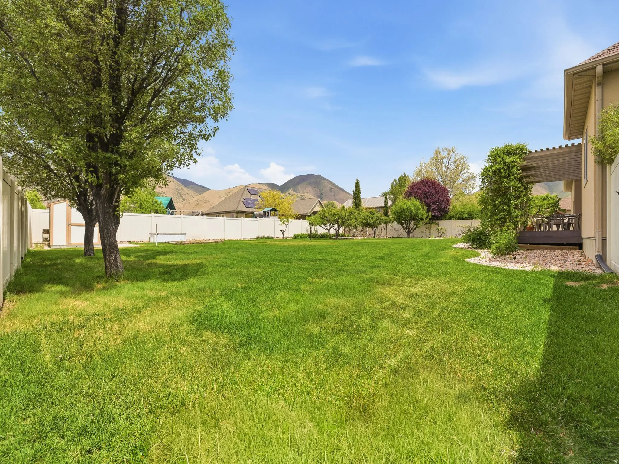 Expansive backyard featuring a lush lawn, mature trees, white vinyl fencing, and an elevated patio with a pergola