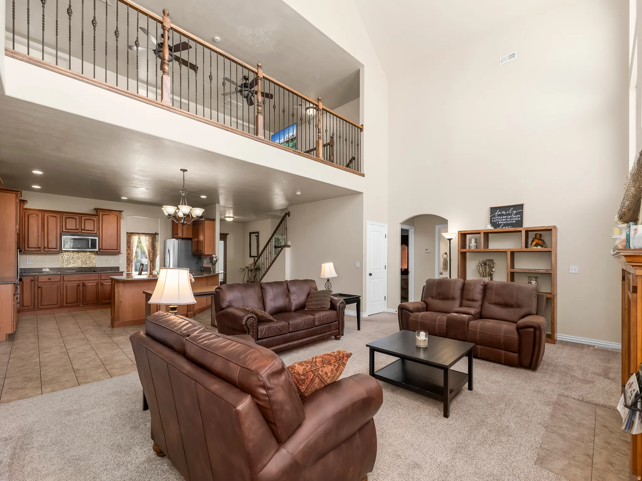 Vaulted living space featuring an upper-level balcony with wrought iron spindles, a visible kitchen with wood cabinetry and tile flooring, and a carpeted living area