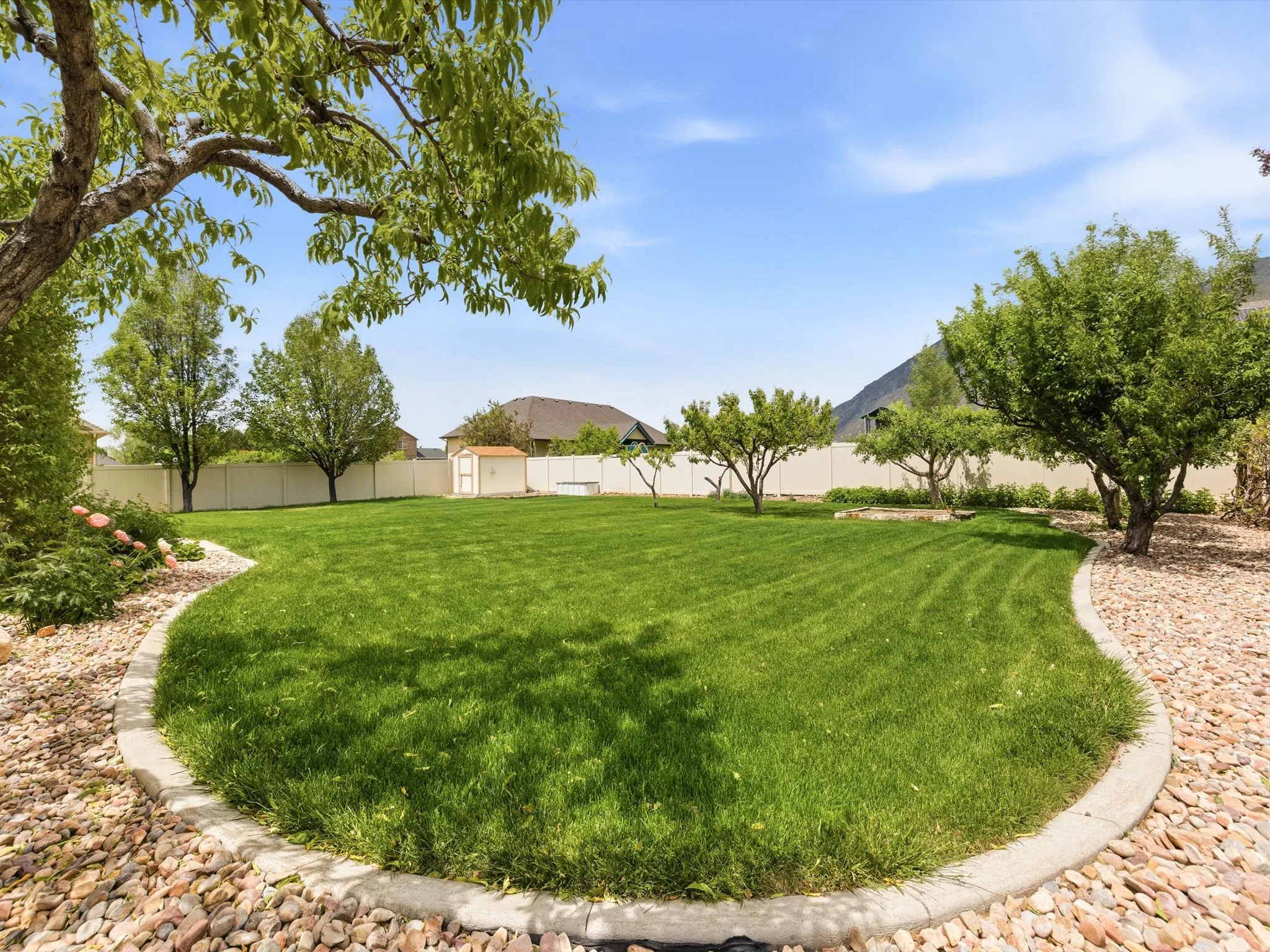 Expansive backyard featuring a lush lawn, mature trees, decorative rock landscaping, a white privacy fence, and an exterior storage shed