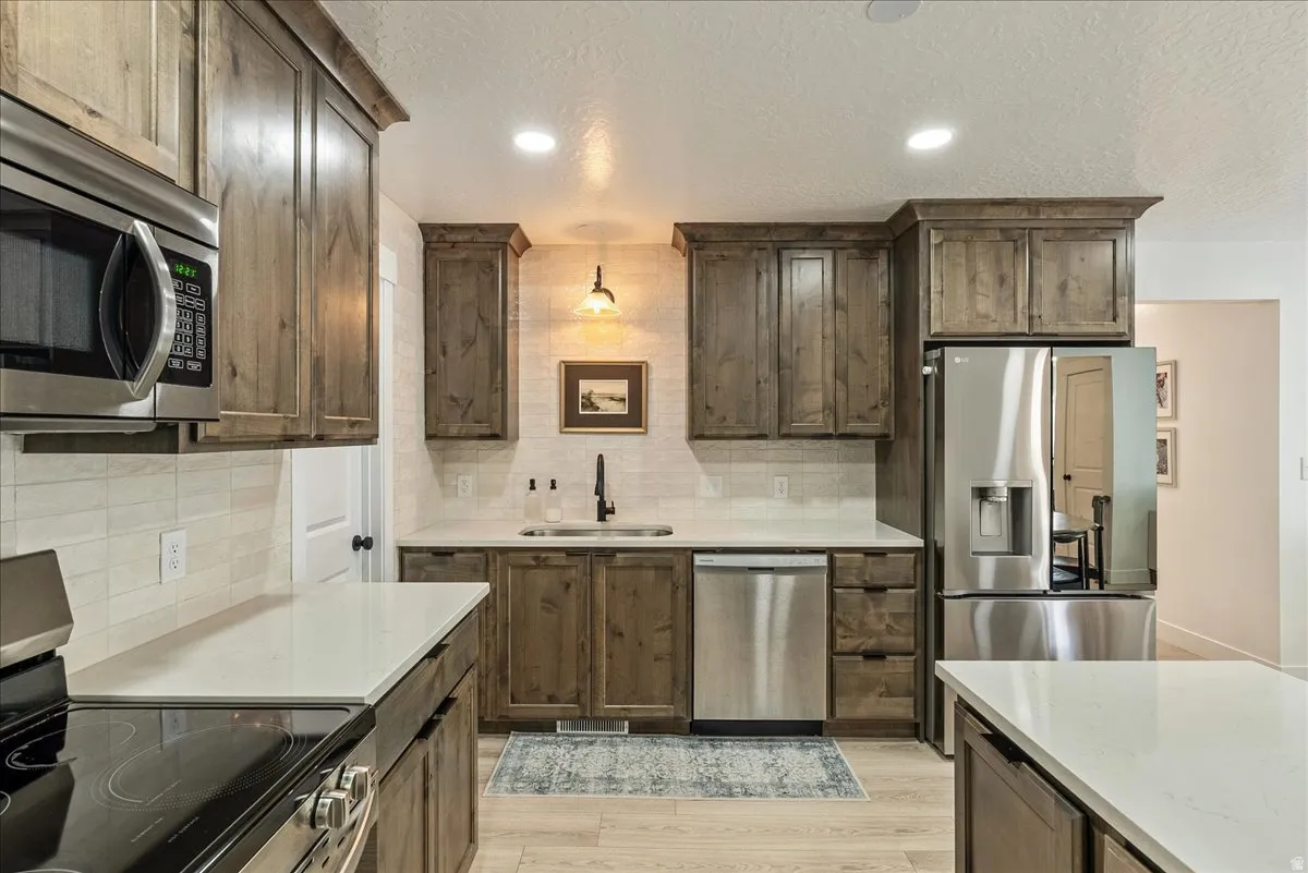 Kitchen featuring wood-finish cabinetry, stainless steel appliances, light-toned countertops, tile backsplash, and light wood-finish flooring