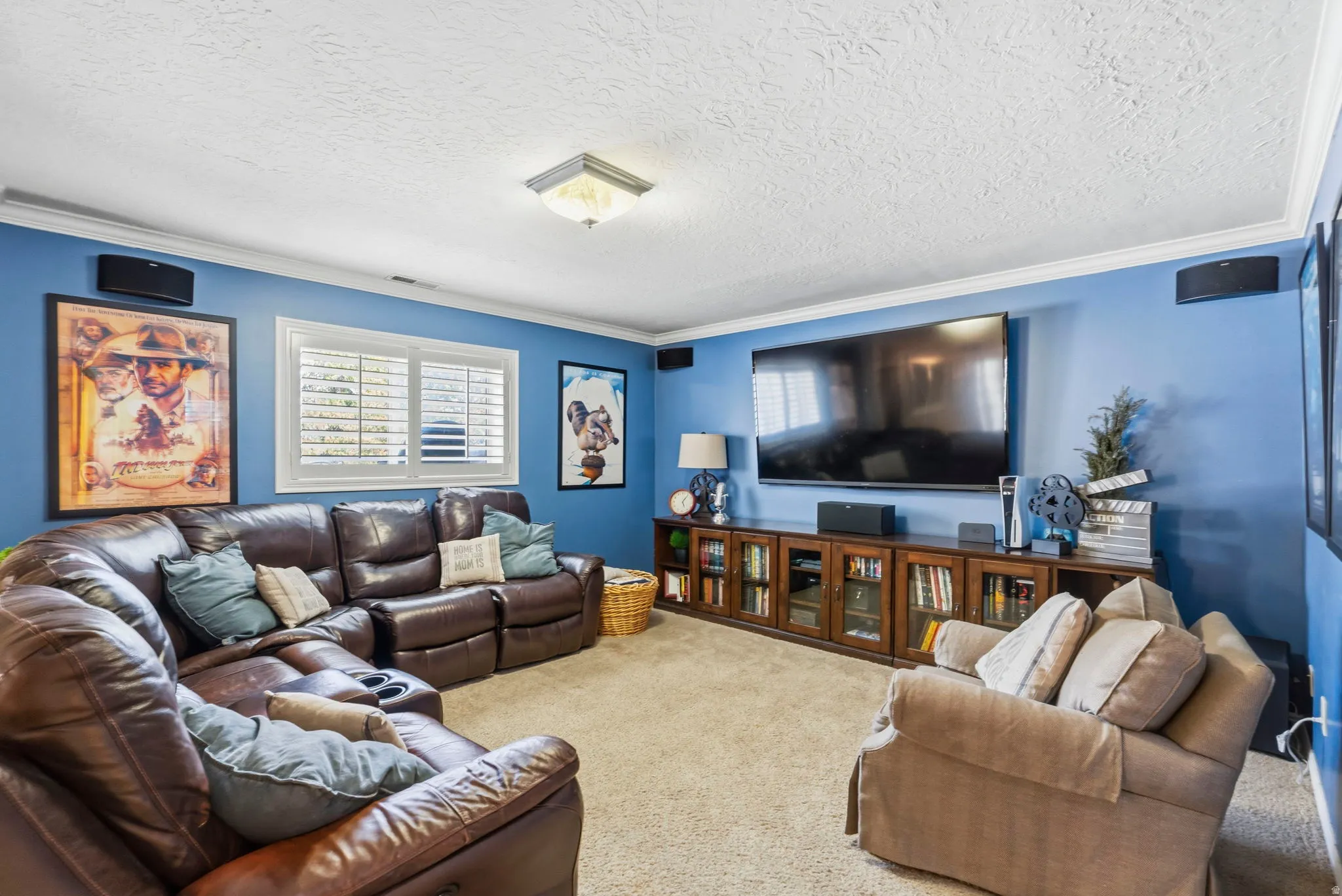 Carpeted recreation room featuring crown molding, a large built-in media console with glass-front cabinetry, multiple wall-mounted speakers, and white window shutters