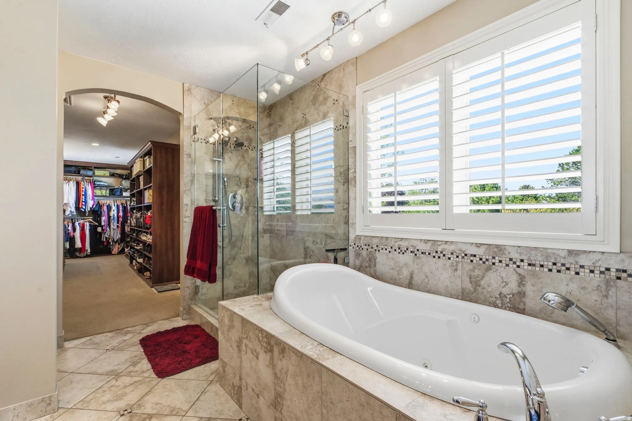 Spacious bathroom featuring an integrated soaking tub, clear glass shower enclosure, stone-finish tile surround, and plantation shutters